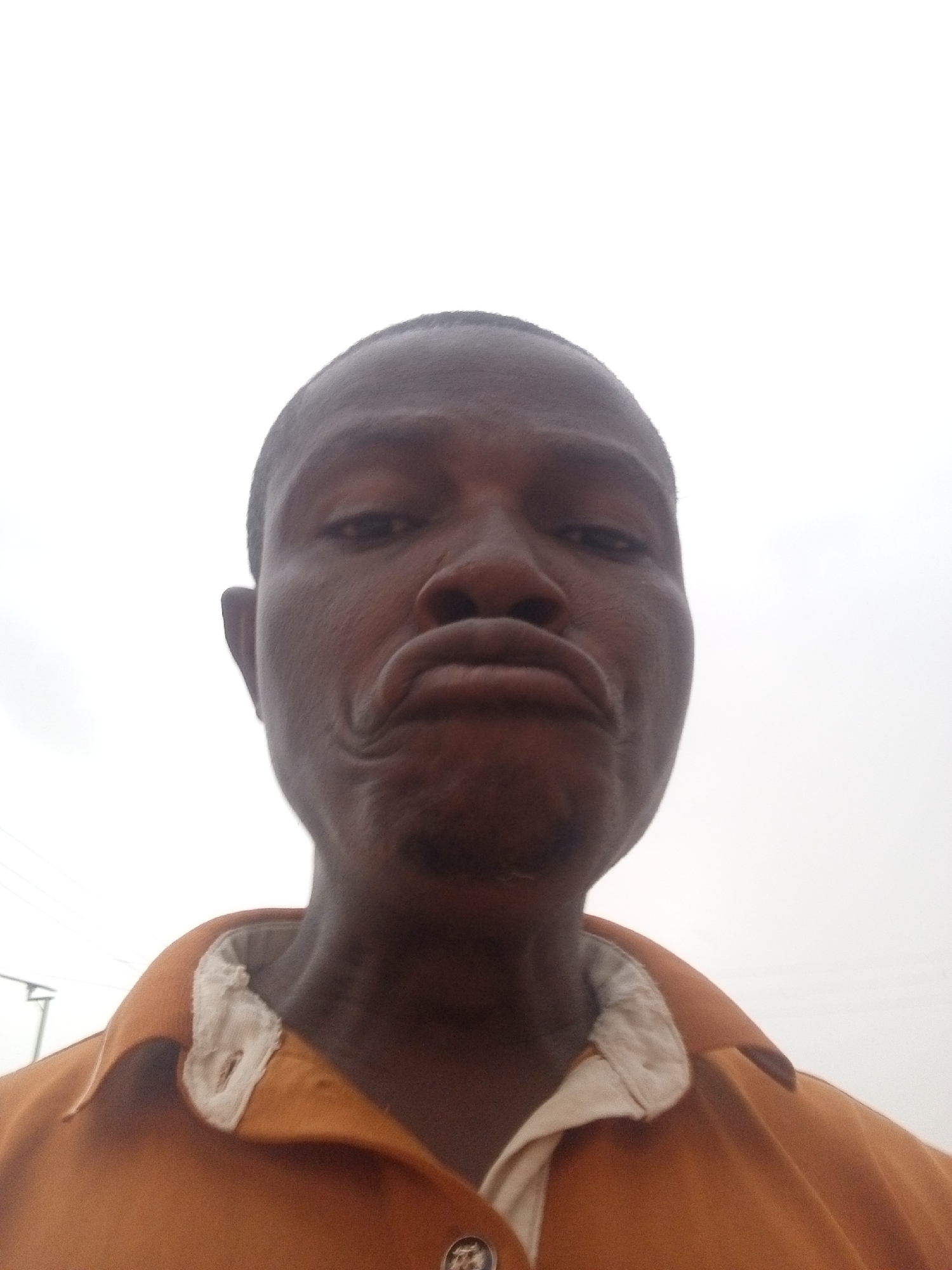 A low-angle close-up photograph depicts a Black man with a shaved head, pursed lips, and a somewhat grim expression. He is wearing an orange collared shirt with white fabric visible at the collar, secured with a button. The background is a light gray sky with some blurred, unidentified structures in the far left. The location appears to be outdoors in an urban area. The city is Unknown, Unknown.