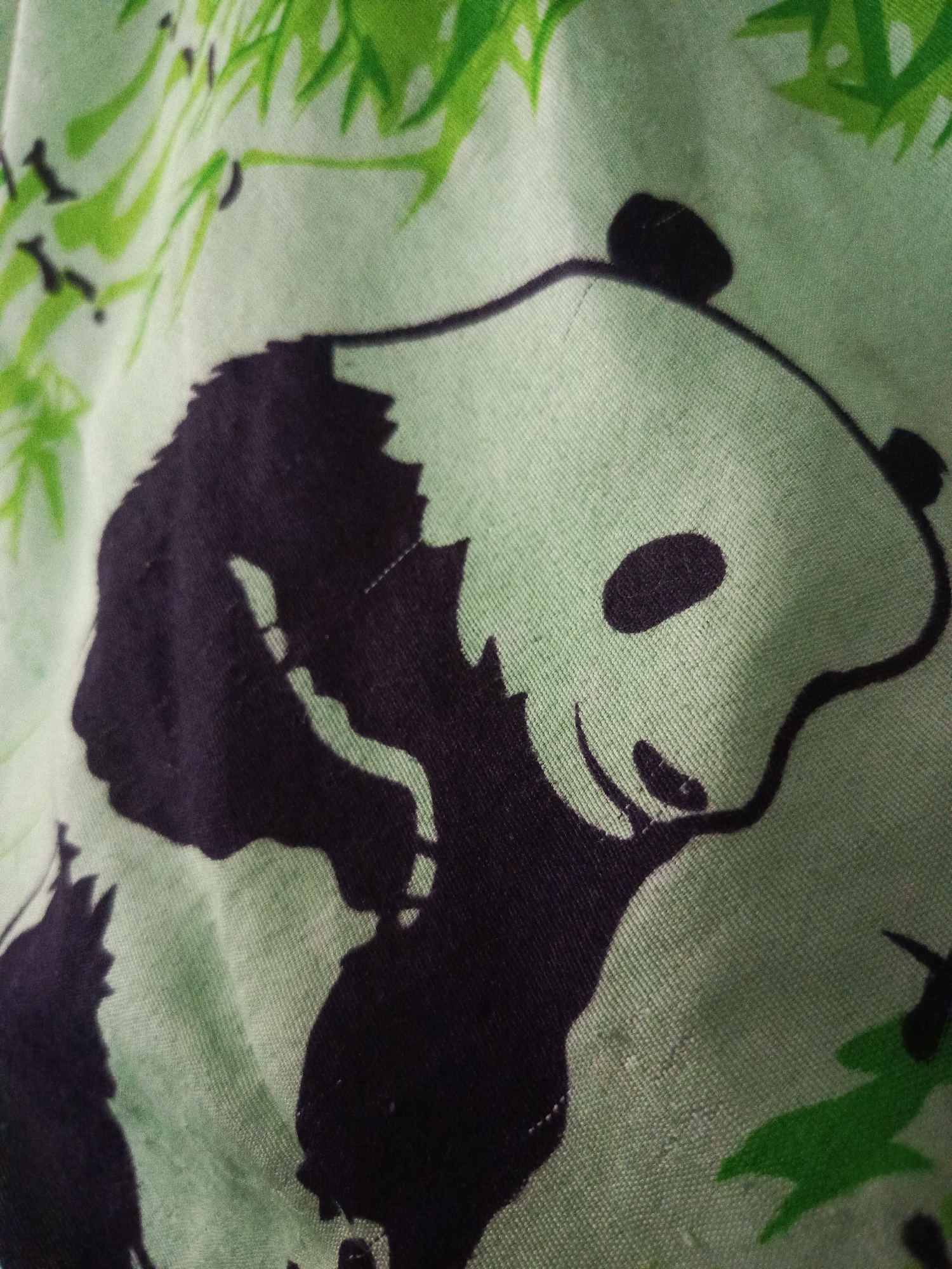 The image is a close-up, top-down view of a light green fabric with a repeating pattern of pandas and bamboo leaves. The pandas are black and white, with large black eye patches and a smiling expression. The bamboo leaves are a vibrant green. The fabric appears to be cotton or a similar textile, with a visible weave texture. There is no indication of the location within the city of Irbeyskoye, Russia, or any other environmental details.