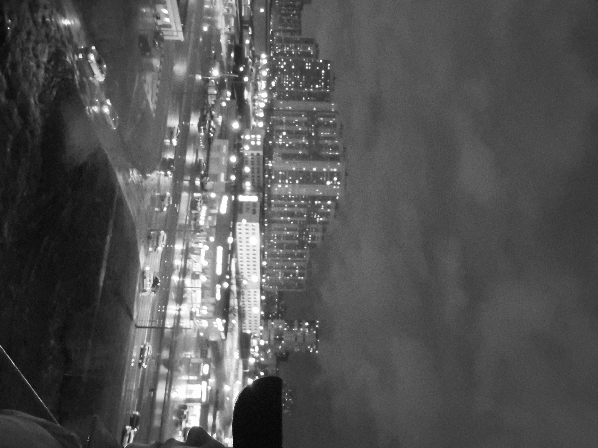 The black and white image appears to show a city scene at night, possibly viewed from an elevated position. A body of water is visible on the left, with reflections suggesting possible rain or wet conditions. A street and buildings with lighted windows stretch across the image. The right side features a large, cloudy sky. The perspective is slightly tilted. An object is visible at the bottom of the frame, obstructing the complete view of the cityscape.