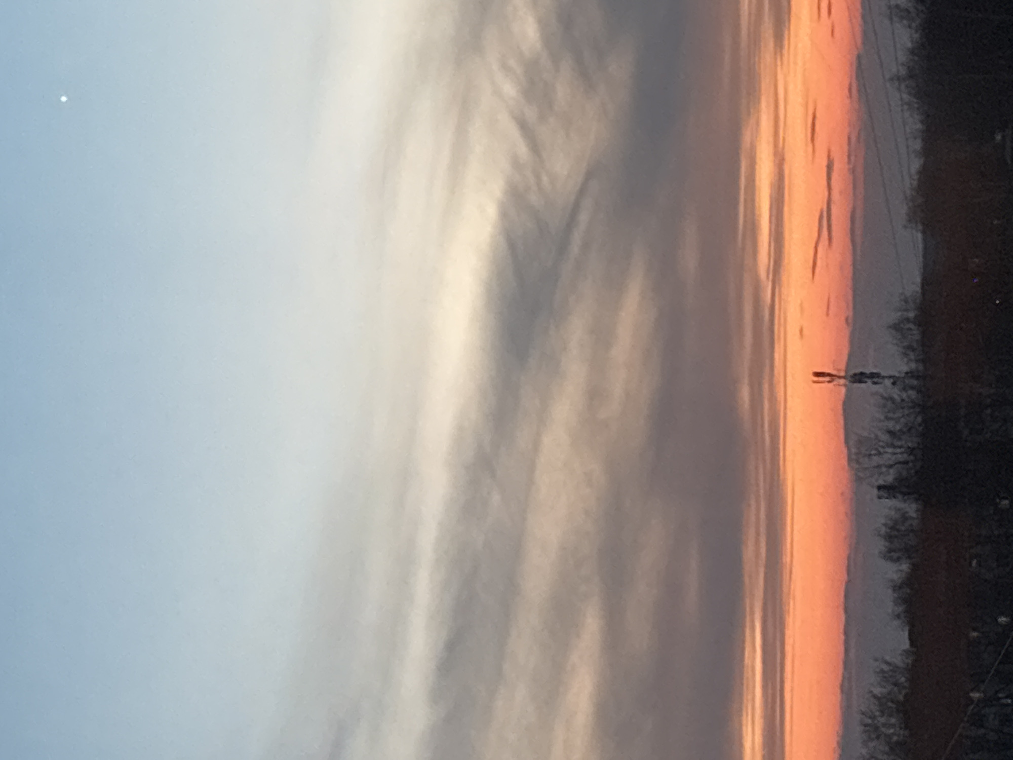 The image shows a view of the sky at sunset, likely taken in the city of Chaikovsky, Russia. The sky transitions from blue on the left side to orange on the right, with some light clouds. There is a small white object in the blue part of the sky, potentially a star or a distant light. The lower right portion of the image shows trees, power lines, and a power pole.