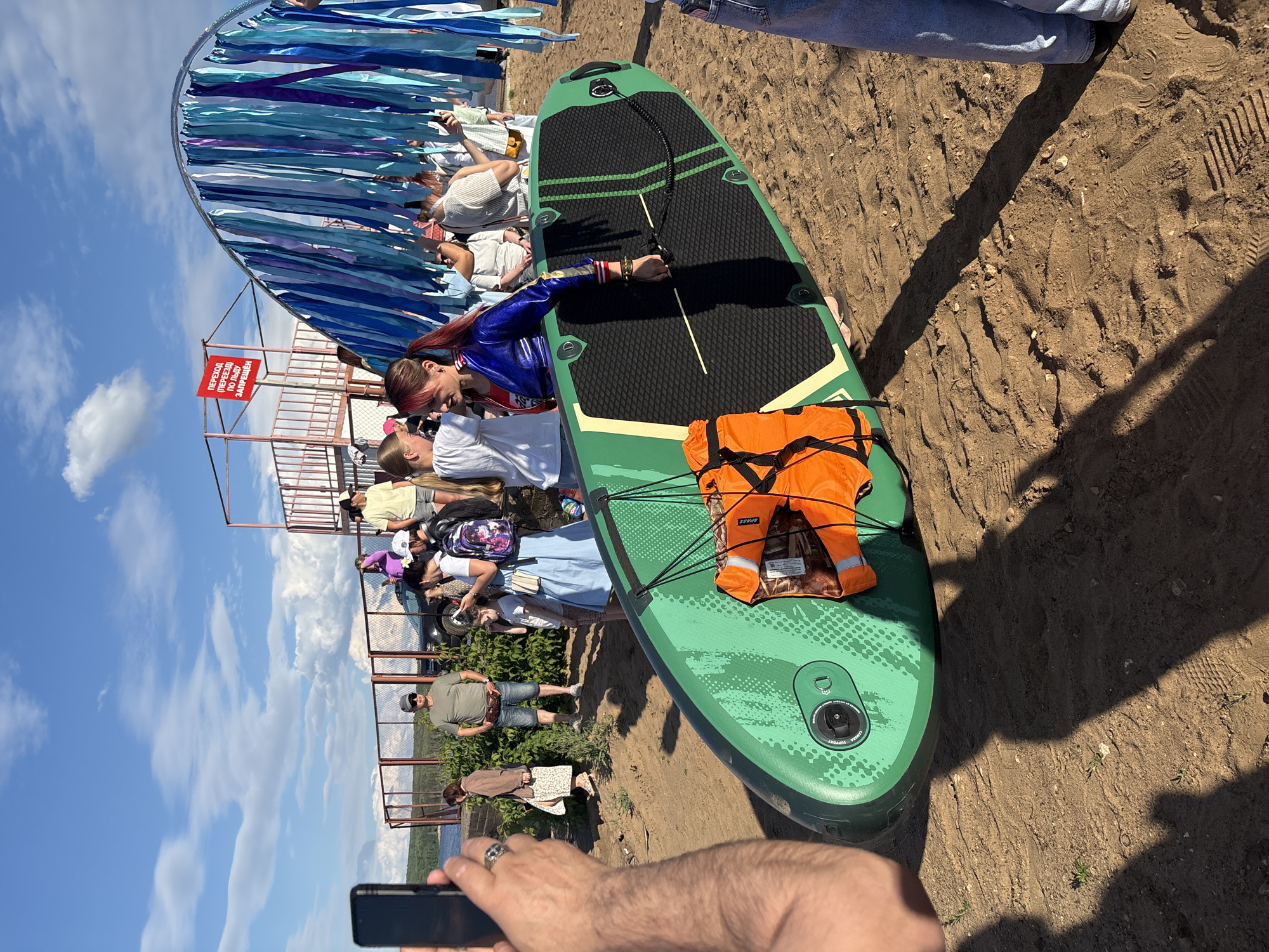 In Chaikovsky, Russia, on a sandy area near a body of water, a group of people are gathered under a bright, cloudy sky. In the foreground, a person is holding a smartphone, seemingly taking a picture. A green paddleboard with an orange life vest strapped to it lies on the sand. A woman in a blue metallic jacket and red top is holding the paddleboard. In the background, several people are standing and interacting, with a fence and some foliage behind them. A red sign with Russian text is visible on a metal structure. The overall scene suggests an outdoor recreational event or gathering.