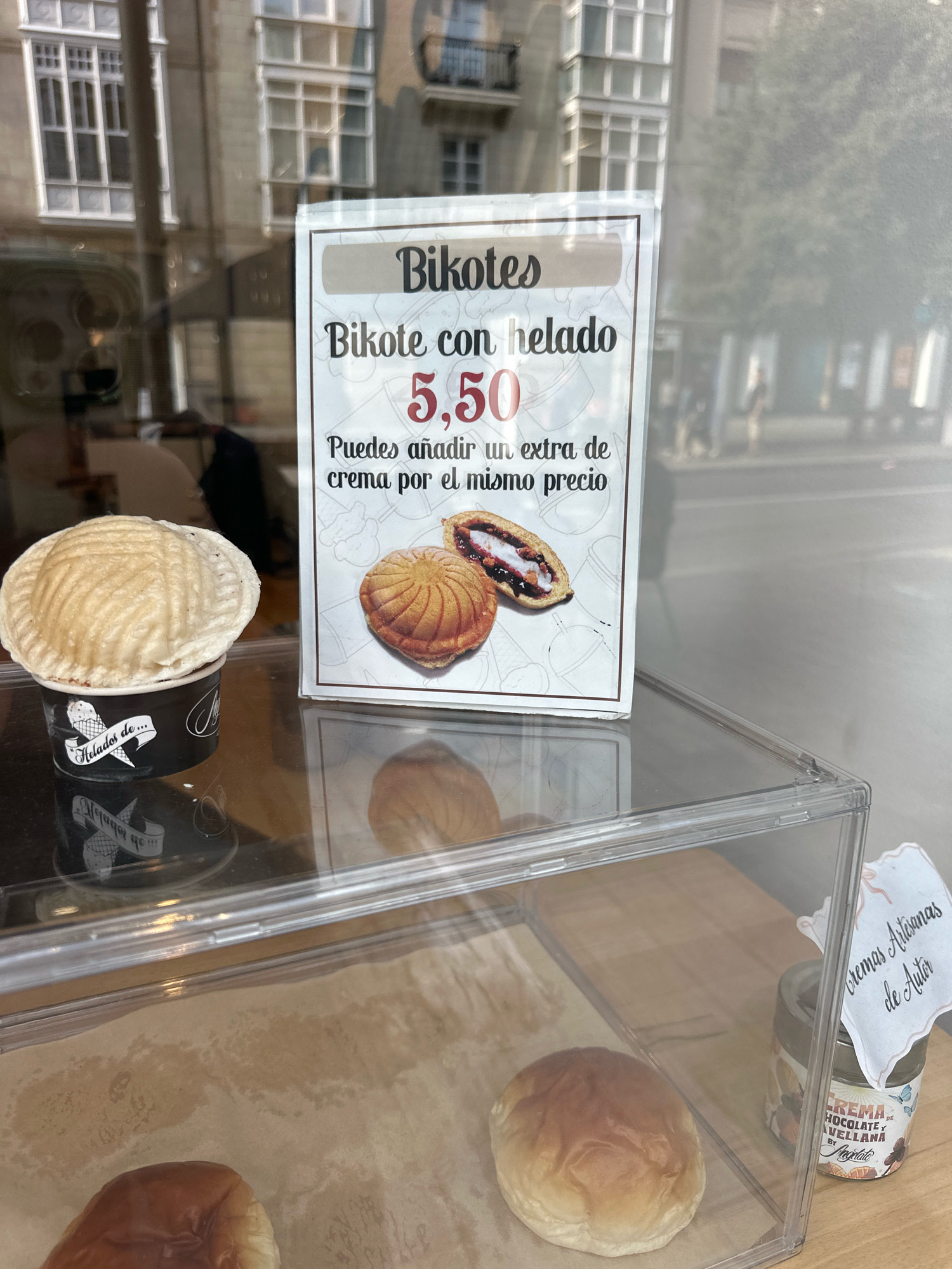 The image captures a display in the window of a bakery or ice cream shop in Vitoria-Gasteiz, Spain. The focal point is a sign advertising "Bikotes con helado" (Bikotes with ice cream) for 5.50. The sign features an illustration of a bikote, a type of sweet bread, split open to reveal a filling, and an ice cream cup. To the left of the sign, a bikote topped with a scoop of ice cream sits in a branded cup labeled "Helados de...". The display case below shows several more bikotes and a jar of "Crema Chocolate y Avellana" (Chocolate and Hazelnut Cream). The background reflects a street scene with buildings and pedestrians, characteristic of a commercial area in Vitoria-Gasteiz. The lighting suggests it is daytime, likely outdoors, with sunlight illuminating the display and casting reflections.