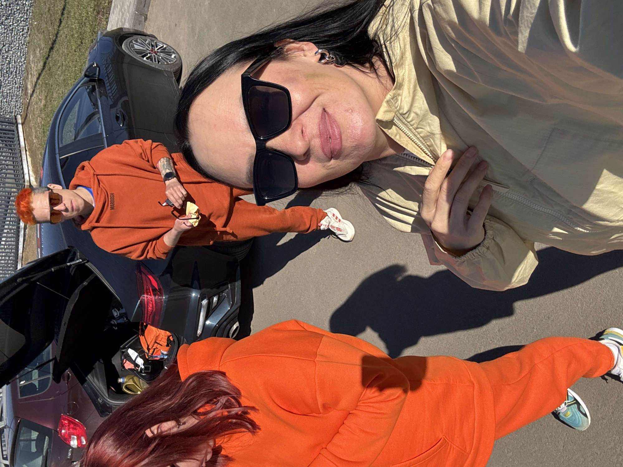 The image depicts three individuals next to parked cars. One person, wearing a beige jacket and sunglasses, appears to be taking the selfie. Another person is clad in an orange hooded sweatshirt and trousers, holding sunglasses and a small cloth. The third individual is wearing matching orange clothing and red hair. Car trunks are open. The location is outside in daylight, with elements suggesting an urban environment, though without specific identifying features pointing to Votkinsk, Russia.