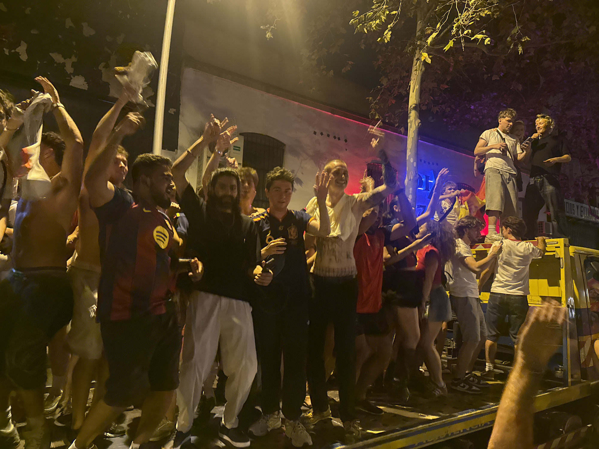 This image depicts a gathering of people at night, likely celebrating a victory or event, on a street in Barcelona, Spain. The group is standing on the back of a yellow flatbed truck, some with their arms raised in excitement. Several individuals are wearing sports jerseys, with one prominently displaying the FC Barcelona colors and crest. One person is holding a microphone, possibly addressing the crowd. The background shows a building with lights and a tree, suggesting an urban environment. The overall atmosphere appears festive and energetic.