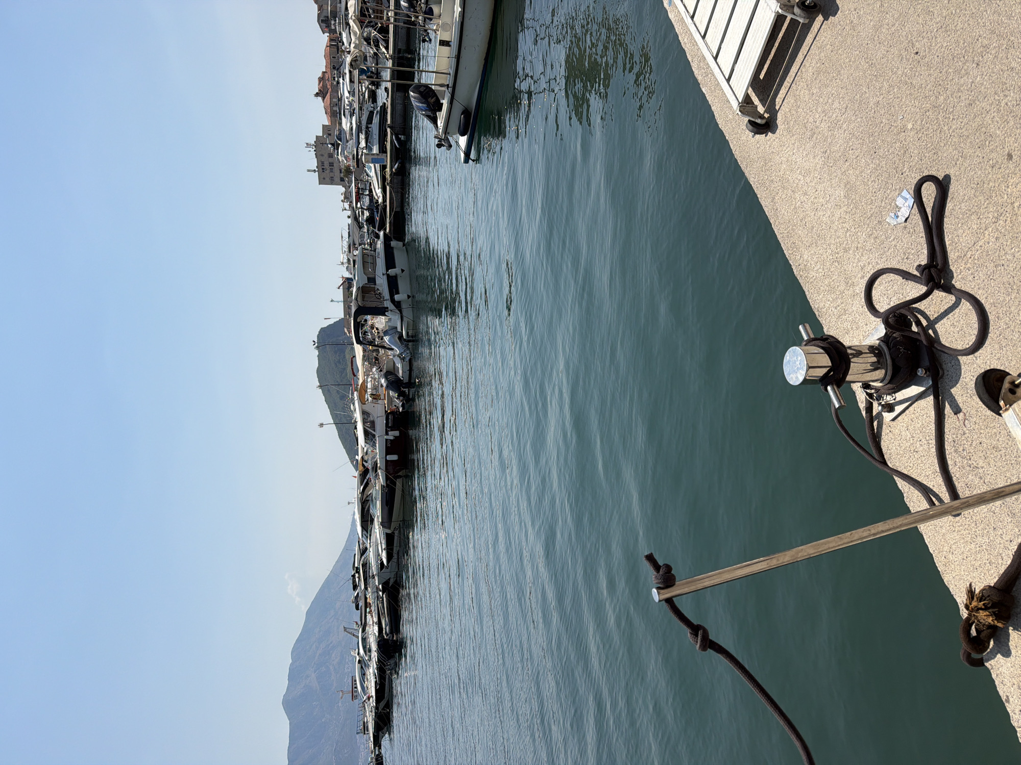 A bright, sunny day in a marina in Budva, Montenegro, is captured from a low angle. The foreground features a concrete pier with a silver bollard and thick black ropes tied to it. A metal support pole with a rope attached stands upright in the foreground, extending towards the water. Beyond the pier, a body of calm, blue-green water is visible, reflecting the clear sky. A line of various motorboats and yachts are moored in the mid-ground, most of them white with dark accents. In the background, a mountainous landscape with a prominent peak dominates the left side of the frame, while buildings and more boats are visible on the right. The sky is a clear, pale blue. There are no people visible in the image.