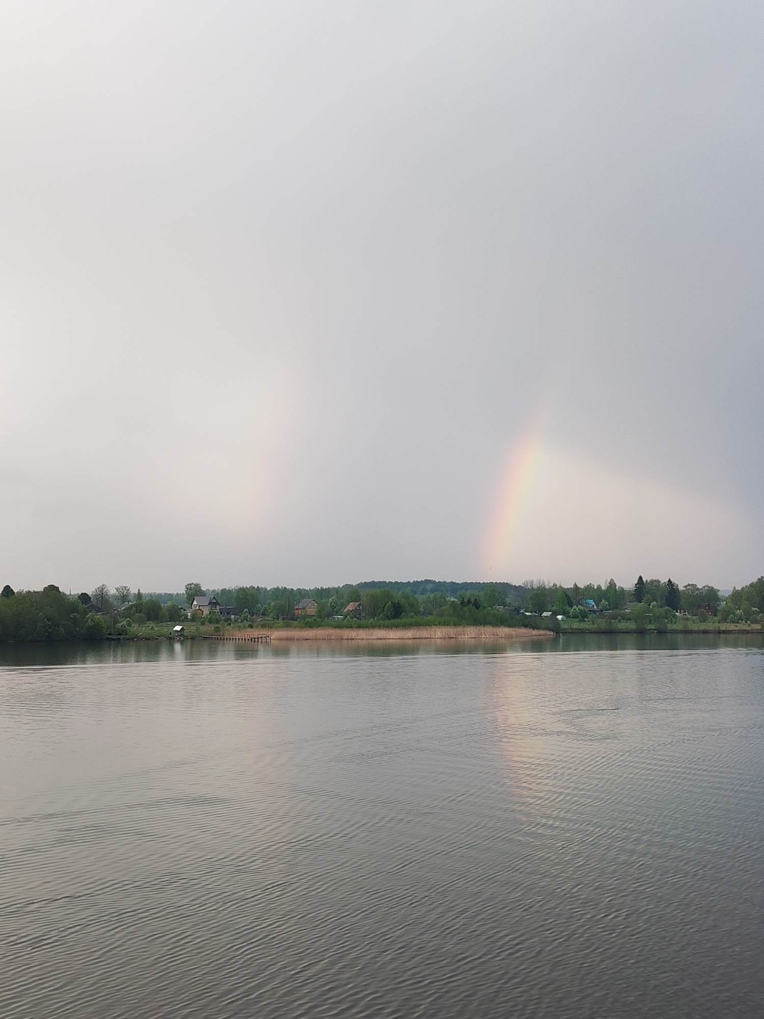 The image depicts a tranquil water scene, likely a lake or wide river, in the vicinity of Анхимово, Russia. The water has gentle ripples across its surface. The shoreline is lined with a mix of trees and reeds, suggesting a natural or semi-natural environment. A few houses are visible among the trees, indicating the presence of a small settlement or village. A distinct rainbow is present in the sky, its arc partially visible. A reflection of the rainbow appears mirrored in the water. The sky is overcast, with a muted, gray tone.