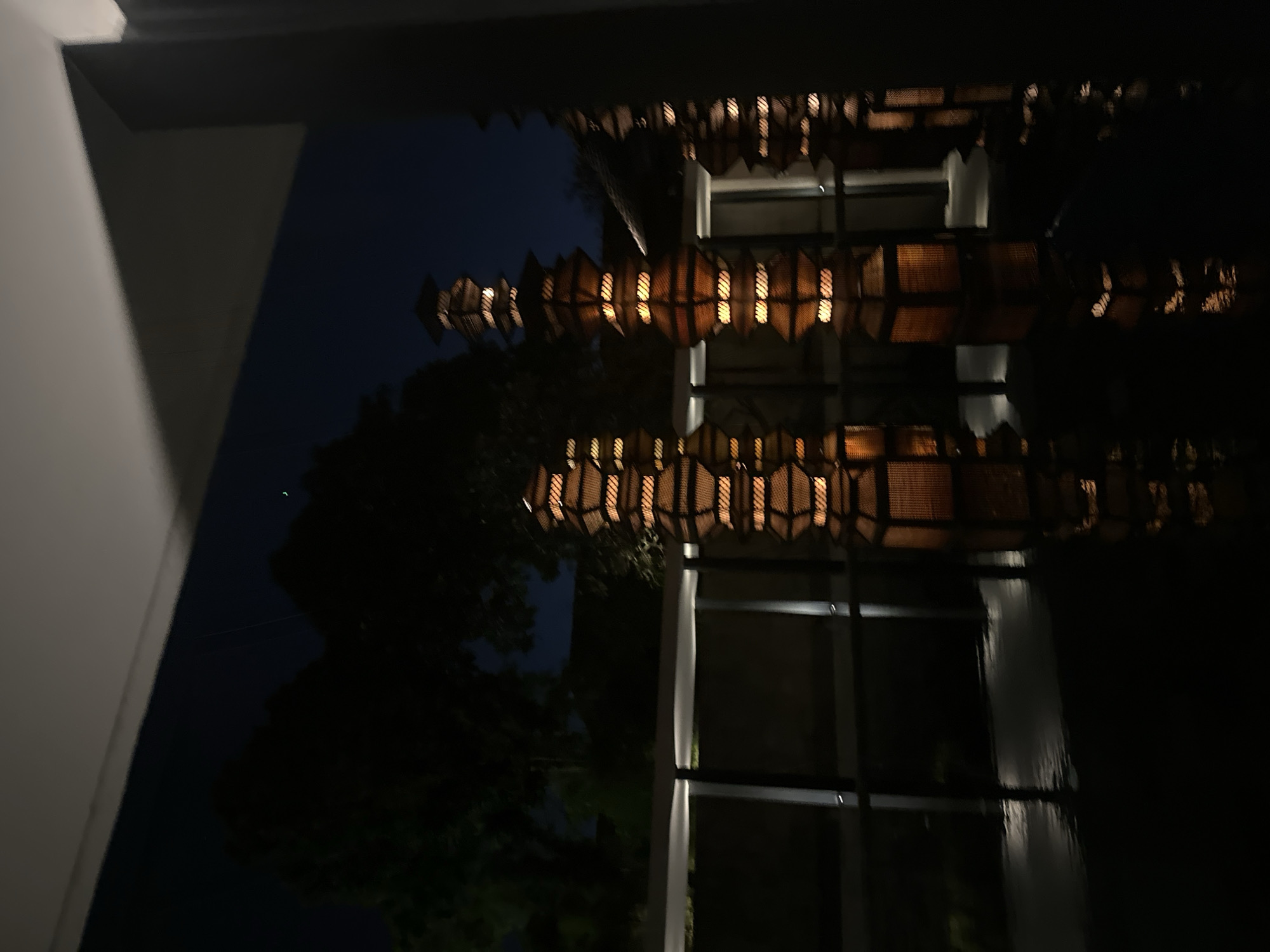 The image captures an outdoor scene at night, likely in Kuta, Indonesia. The primary subjects are several decorative lanterns with a woven texture, illuminated from within, casting a warm glow. These lanterns are suspended from an overhang, creating a repeating pattern of light and shadow.
The setting appears to be modern, with hints of a building's architecture visible, including large windows or glass panels that reflect the lanterns. Trees are silhouetted against the dark night sky, suggesting a natural element within the urban or resort environment. The overall mood is serene and atmospheric, enhanced by the low light and the gentle illumination of the lanterns.
There are no people visible in the frame. The time of day is clearly night, indicated by the darkness and the artificial lighting. The weather is not explicitly discernible, but the calm reflections suggest still conditions. The location context of Kuta, Indonesia, aligns with the tropical foliage and the style of the lanterns, often found in resorts or hospitality venues in such areas. No text is visible in the image.