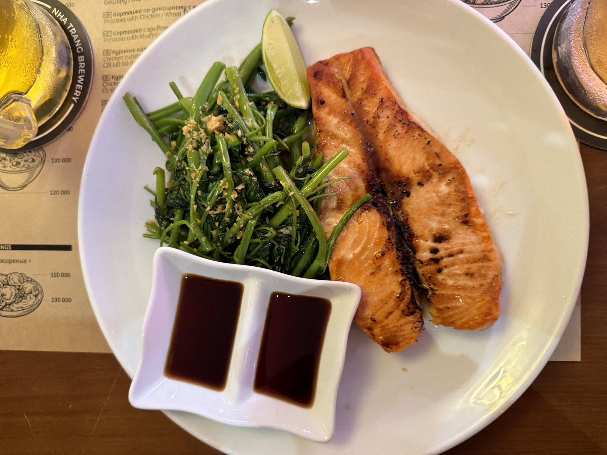 A close-up shot captures a white plate of food on a wooden table. The plate contains two pieces of grilled salmon, a mound of stir-fried green vegetables, a lime wedge, and a small white dish with two compartments filled with dark sauce, likely soy sauce. The background reveals a menu with Vietnamese text and prices, a coaster with "NHA TRANG BREWERY" printed on it, and a glass of beer. The lighting suggests an indoor setting, possibly a restaurant in Nha Trang Ward, Vietnam. No people are visible in the image.