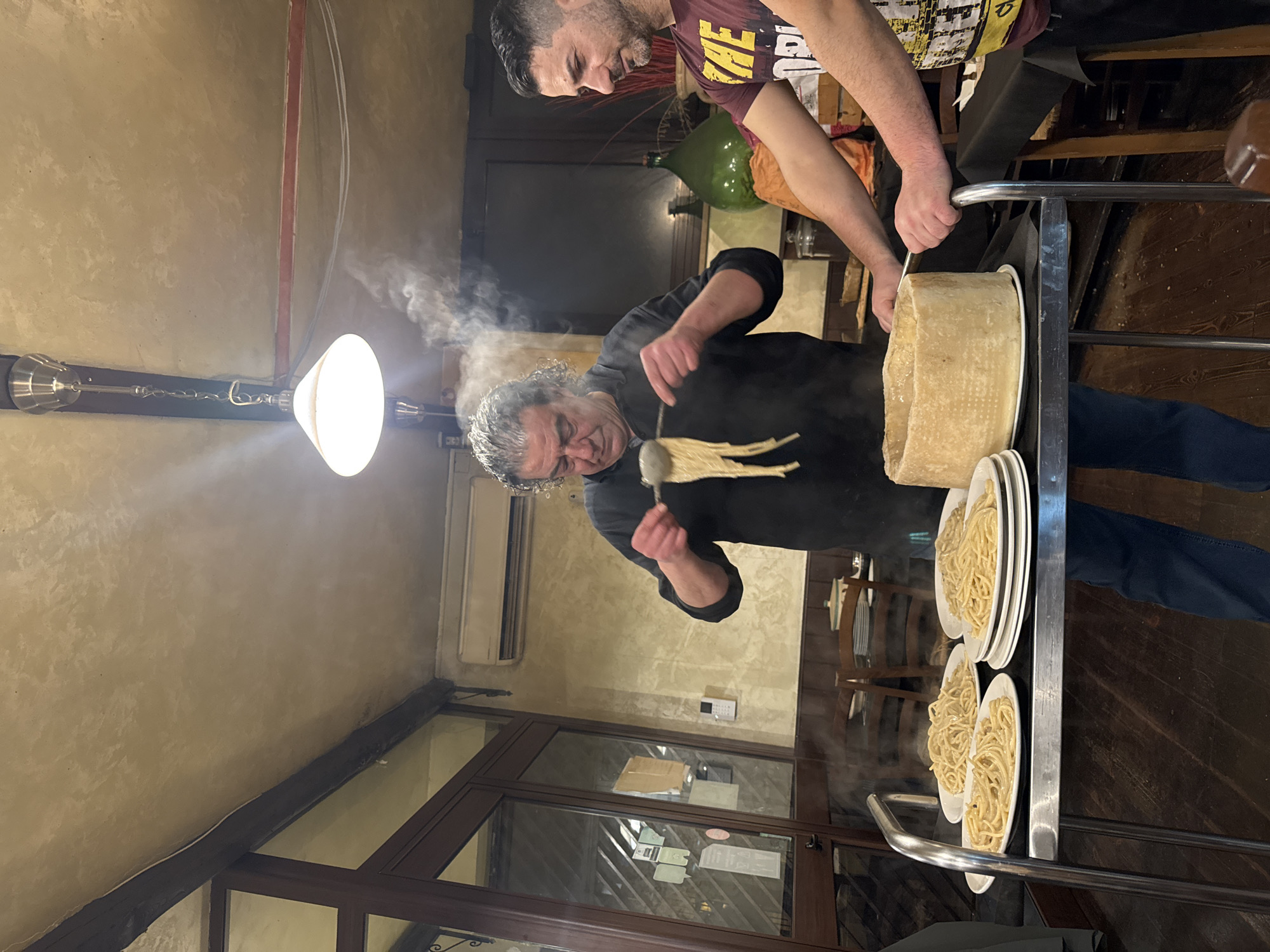 The image depicts an indoor scene within a traditional Roman trattoria in Rome, Italy, during what appears to be evening service. Two adult males are centered around a metal-framed service cart, actively engaged in preparing and serving pasta.
The man on the left, with curly grey hair and wearing a black long-sleeved shirt and blue jeans, is the primary preparer. He holds a metal serving utensil, from which strands of freshly prepared pasta (likely spaghetti or bucatini) are suspended, emitting steam. His gaze is directed downwards at the pasta.
On the right, a younger man with short dark hair and a beard, wearing a maroon t-shirt with yellow lettering, stands alongside the cart. He rests his right hand on the cart's railing and his left hand near a large, hollowed-out wheel of aged cheese (likely Pecorino Romano or Parmesan), which functions as a mixing vessel on the cart. He appears to be observing or assisting.
The service cart holds multiple white plates, each containing a portion of pasta, arranged around the cheese wheel. Steam visibly rises from the pasta on the plates and from the area of the cheese wheel.
The background reveals the restaurant's interior: textured, light-colored walls with dark wooden wainscoting, a dark wooden floor, and wooden tables and chairs indicating dining areas. An air conditioning unit is mounted on the left wall, above a dark-framed glass door or window. A large green glass demijohn is visible on a counter in the background to the right. A prominent white, cone-shaped pendant light fixture hangs directly above the service cart, illuminating the activity. The ceiling is light-colored and textured, featuring exposed dark wooden beams.