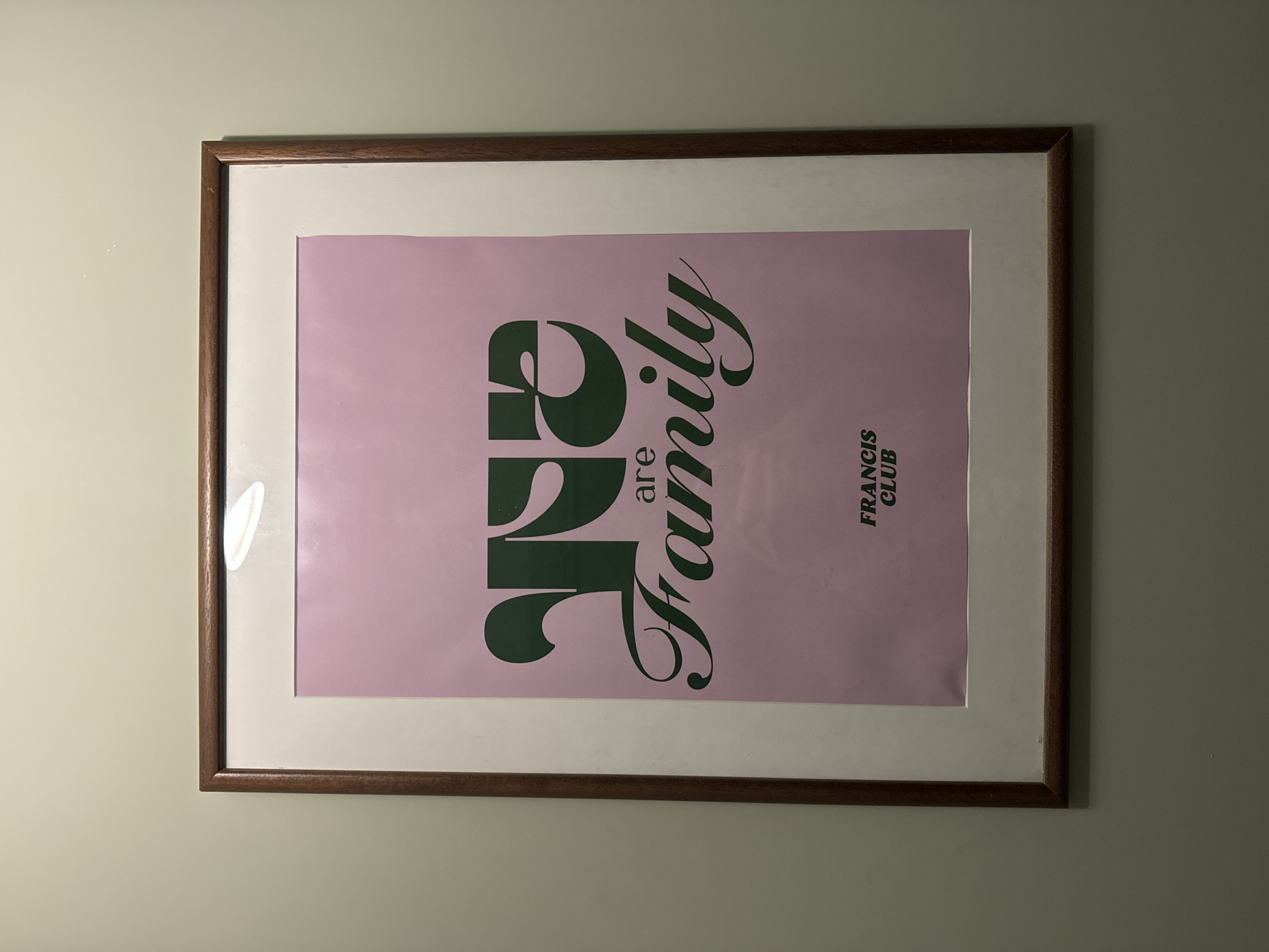 A framed pink poster hangs on a wall. The poster has the words "are Family" in green text, written vertically. The words "FRANCIS CLUB" are on the right side of the poster. The frame is wood with a white mat. The wall is a light green color. The location is unknown; there is no indication of it being in Krasnoyarsk, Russia.