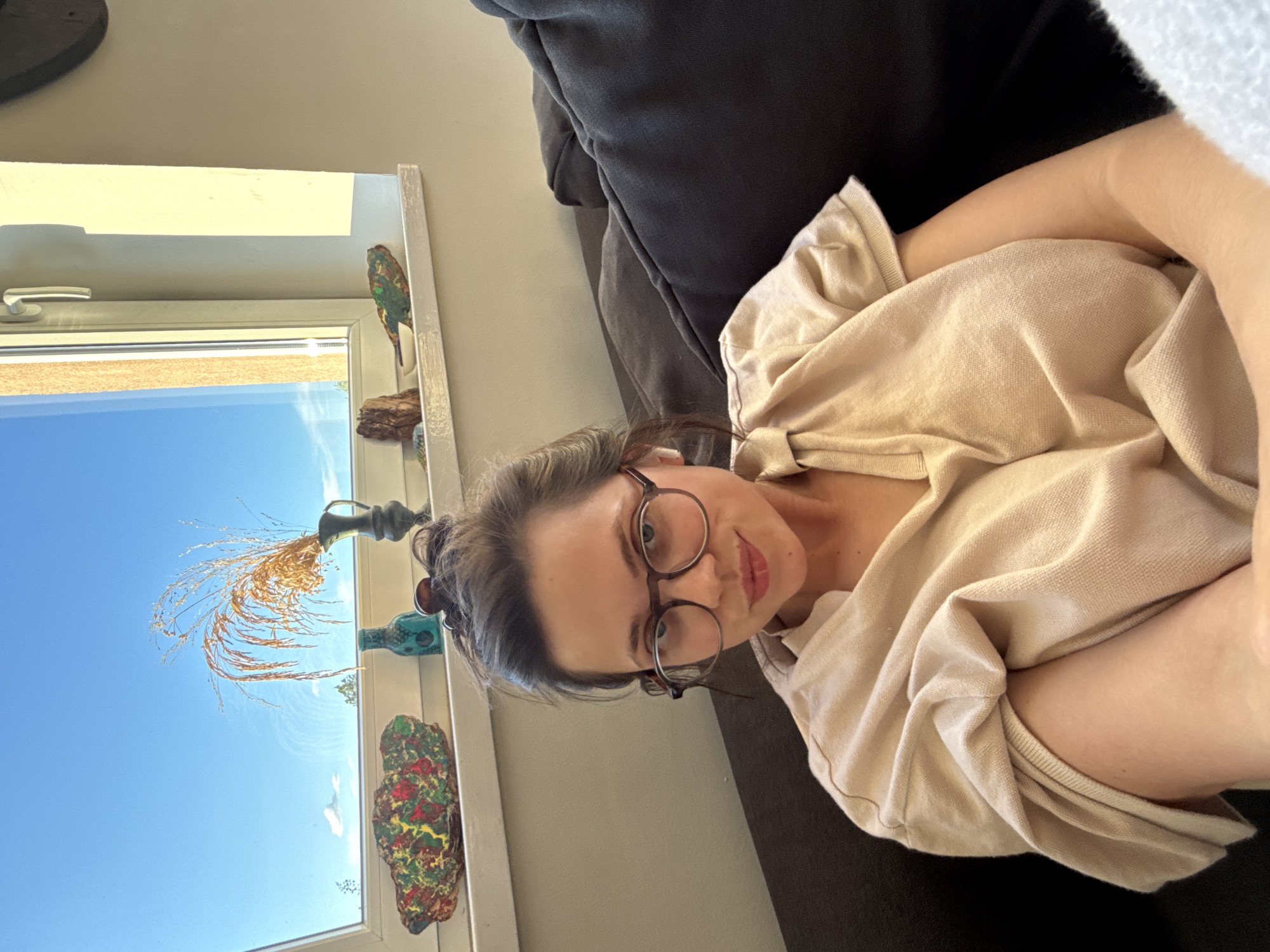 A young woman reclines comfortably on a dark grey or black couch, looking directly at the camera with a calm, slightly smiling expression. She has dark hair styled in a bun, wears round-framed eyeglasses, and is dressed in a light beige or tan long-sleeved top. Her arms are loosely folded.
Behind her, a large white-framed window reveals a clear, bright blue sky, suggesting it is daytime and sunny. Through the window, a light brown/orange building facade is visible in the upper portion, and some green foliage can be seen in the distance near the horizon. The window ledge, also white, is adorned with several decorative items: a colorful, possibly parrot-shaped figurine, a piece of natural wood, an antique-style dark metal vase holding dried golden ornamental grass or reeds, a small turquoise patterned bottle, and another textured, abstract decorative piece in red, yellow, and green. The surrounding indoor wall is a light, neutral color. No text is visible in the image. The setting is indoors, likely a home, with a location context of Budapest, Hungary.