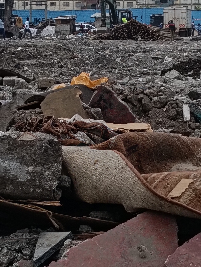 The image depicts a large outdoor demolition or construction site in Nairobi, Kenya, characterized by extensive rubble and debris. In the foreground, fragmented concrete slabs, broken red bricks, and a soiled, rolled-up textile object (likely a rug or carpet in brown and beige tones) are prominent. Pieces of rusty metal and various small debris are interspersed.
The midground features a vast, uneven expanse of dark grey and brown rubble, composed of numerous concrete fragments, stones, and dirt. Several individuals are present: A male figure in a white shirt and dark trousers walks from right to left across the rubble. To the right, a second male figure in a red shirt and dark trousers stands near a white utility truck equipped with a white cargo container. Further back, a substantial pile of cut timber or logs is stacked. Adjacent to this pile, a green and black excavator is positioned, with a person wearing a high-visibility yellow/green vest standing nearby. Scattered makeshift structures, appearing as temporary shelters made of corrugated iron and other salvaged materials, are visible among the debris.
The background reveals a large, prominent blue structure with partially visible white lettering "IKE," possibly indicating "IKEA" or a similar industrial facility. Beyond this structure, other indistinct buildings suggest an urban or industrial setting within Nairobi. The scene indicates ongoing land clearing, demolition activities, or a major waste disposal site.