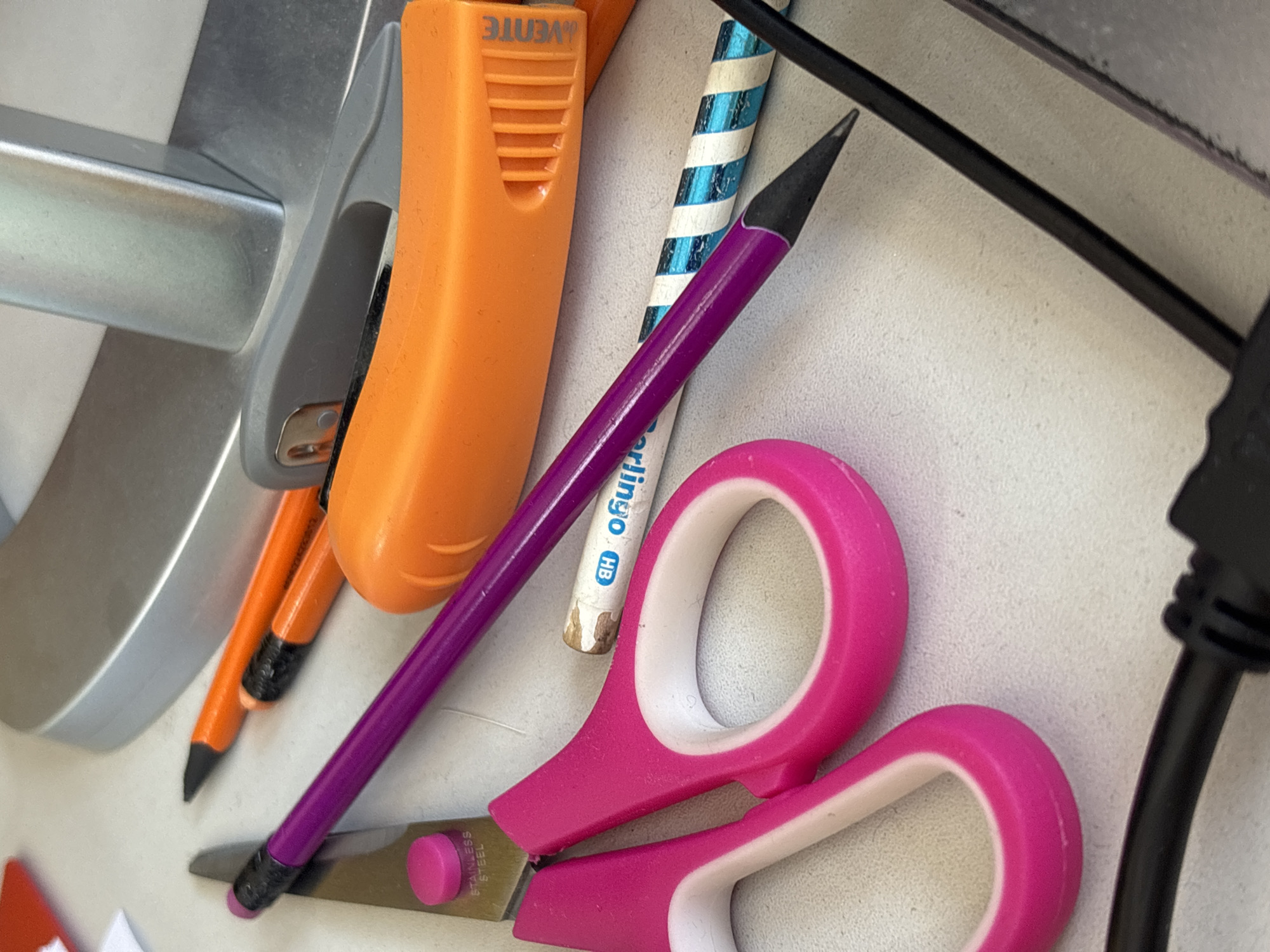 The scene, located in Tayshet, Russia, depicts a static, close-up view of various stationery items and office supplies arranged on a light-colored desk surface. In the foreground, a pair of pink-handled scissors with "STAINLESS STEEL" imprinted on a blade is visible, accompanied by a pink circular stopper. A sharpened purple pencil with a black tip lies across the scissors and other items. Two sharpened orange pencils are situated in the upper-middle section, near a grey stapler with a metallic silver arm. An orange utility knife with a visible "VENTE" logo is positioned to the right of the pencils. Further right, a short white pencil marked "Berlingo HB" and a blue and white striped pencil are present. A large, silver-grey object, likely the base of a monitor, is in the background. A black cable with a dark connector extends into the lower foreground from the right. A small corner of a red object and a piece of white paper are partially visible in the upper left background. No individuals or actions are depicted.
