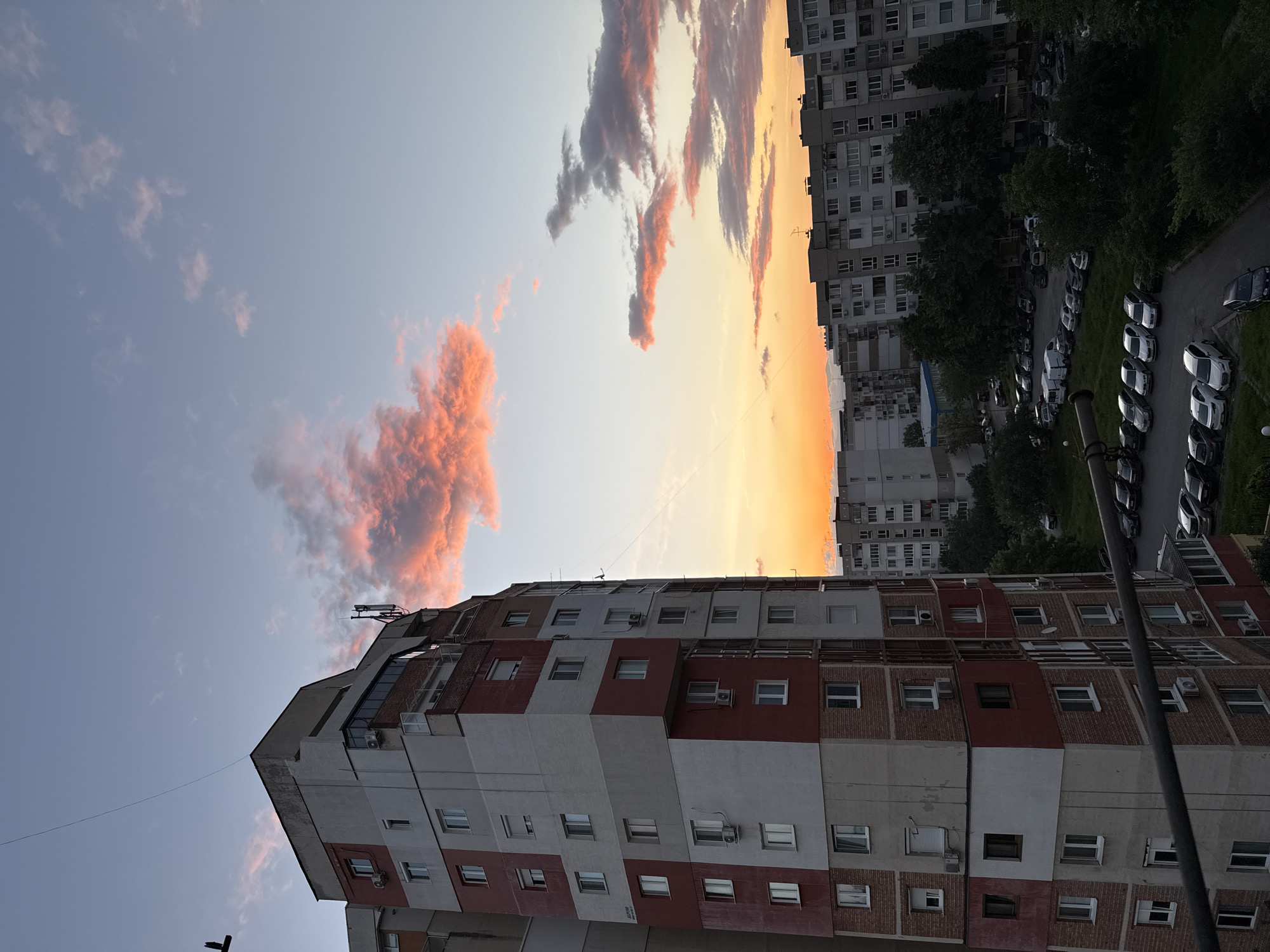 The image shows a view from what appears to be a high-rise building in Sofia, Bulgaria, possibly from a window. The sky displays a sunset gradient with hues of orange and light blue, accented by pinkish clouds. Below the sky, there are multiple buildings, including a tall, multi-story apartment building with alternating bands of red and white. To the right, more buildings and parked cars are visible, partially obscured by trees. A utility pole appears in the lower right corner.