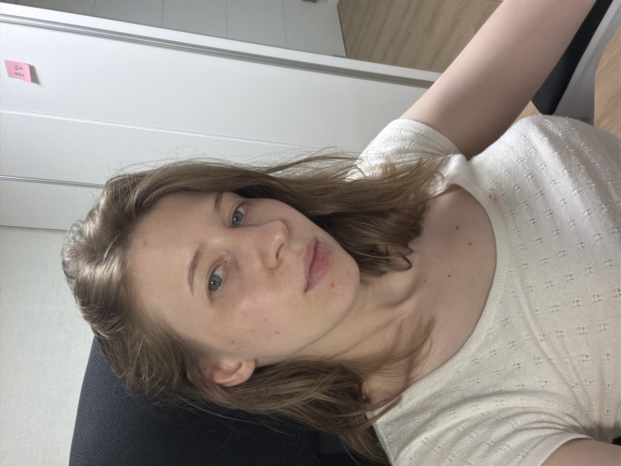 The image shows a close-up, waist-up view of a young woman with light brown hair and blue eyes, seated in a black office chair. She is wearing a white, textured t-shirt with a low neckline. Her skin appears fair with some freckles and slight redness on her cheeks. A small pink sticky note with Korean writing is visible on a white door in the background, suggesting the location is likely an indoor office or room. The room also appears to have light-colored wood flooring. The woman is looking directly at the camera, with a neutral expression. The overall lighting is soft and diffused. The scene is devoid of any notable activity or interaction.