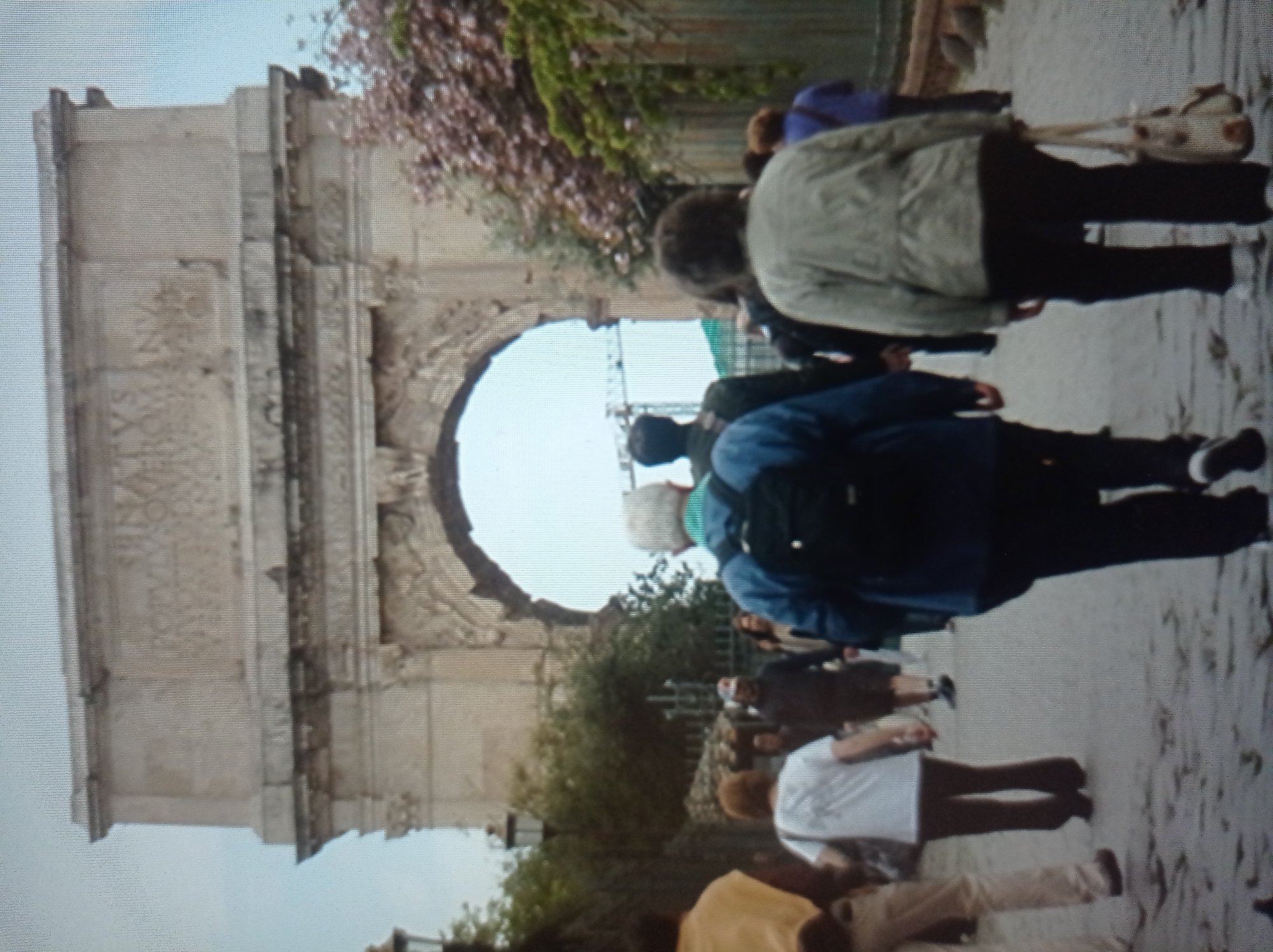 The image shows a view of a stone archway, possibly a historical monument, with people walking through a cobblestone street in front of it. The arch is large and constructed from stone blocks, with ornate carvings visible on its upper facade. Lush green trees and some flowering bushes, with pinkish-red blossoms, are visible on the right side of the arch. Several individuals are walking away from the camera on the street. One person in the foreground is wearing a blue jacket and a backpack. Another person in the middle ground is wearing a light-colored jacket and carrying a light-colored bag. The lighting suggests it is daytime. The image does not provide any information indicating the location is in Mukachevo, Ukraine; it is more likely to be a historical site in a European city.