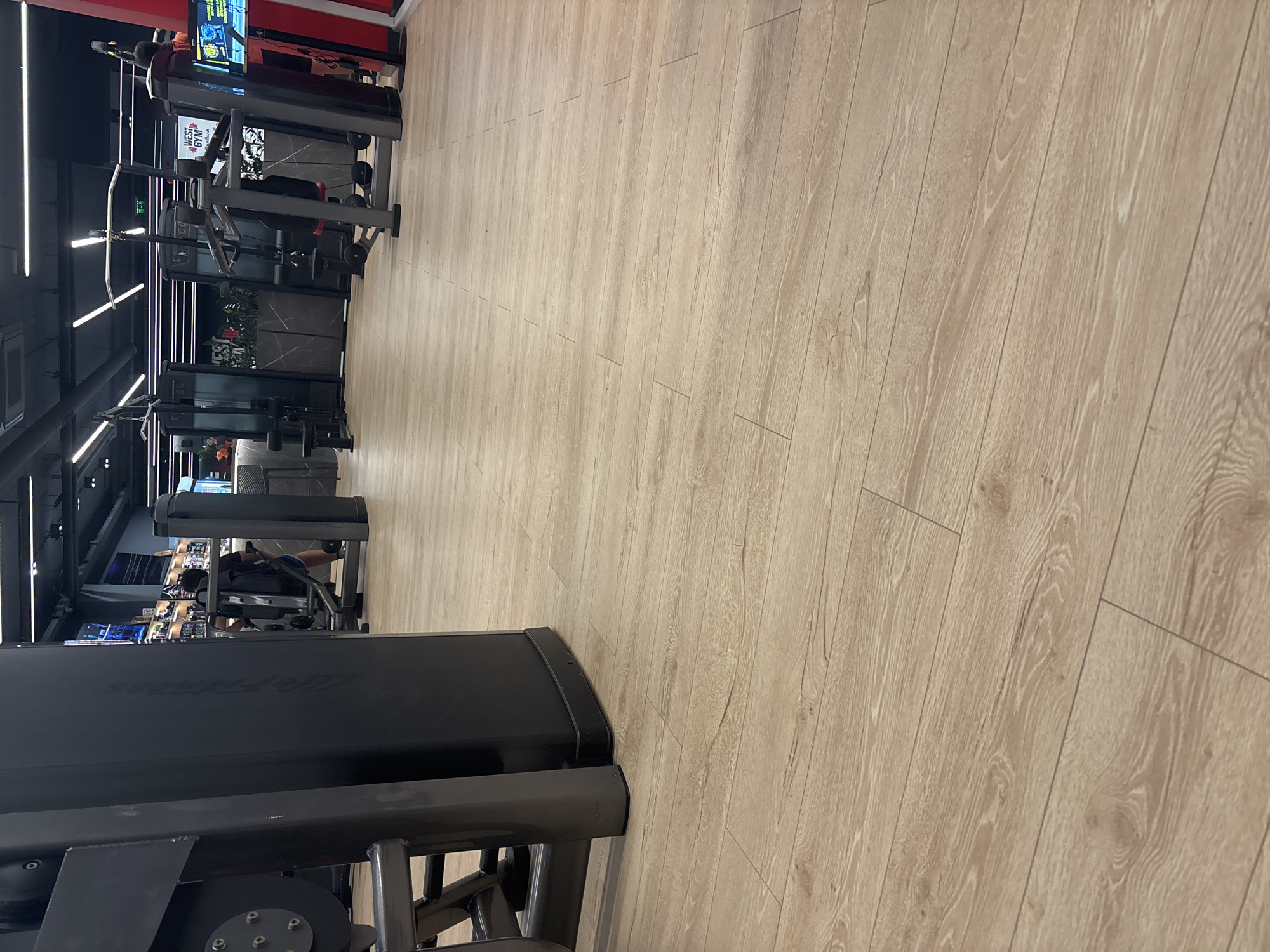 The image depicts the interior of a gym, likely in Sofia, Bulgaria. The foreground features a black "Life Fitness" branded leg press machine. Rows of other gym equipment are visible, including weight machines and treadmills. A few people are using the equipment. The floor has light wood-look flooring. The gym is illuminated by linear lighting fixtures along the ceiling. The setting is a fitness center with standard gym equipment and layout.