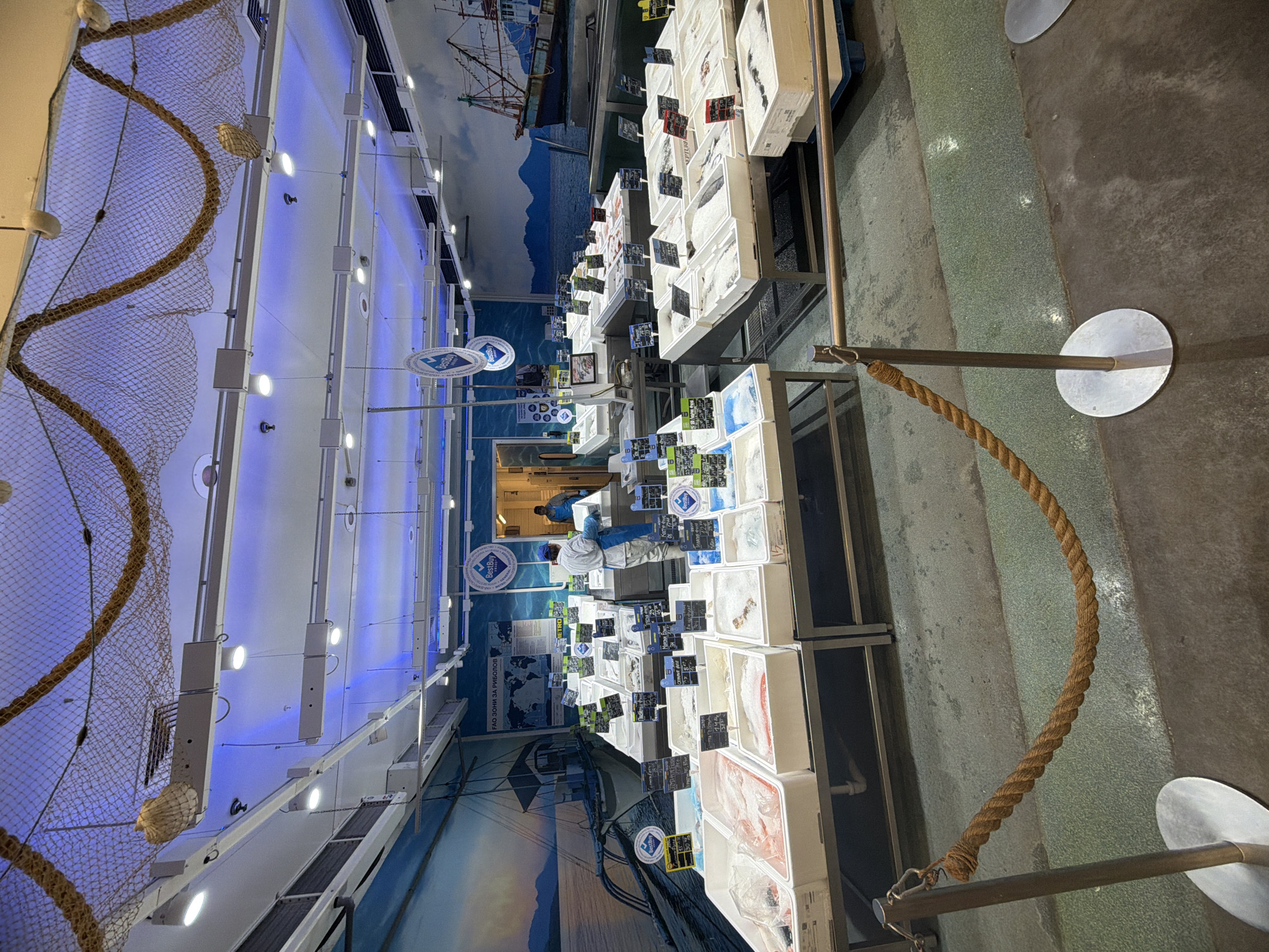 A brightly lit, indoor fish market in Burgas, Bulgaria, features rows of refrigerated display cases stocked with fresh seafood in white containers. Small black price tags are attached to each display. In the background, a mural depicts a seaside scene with boats and mountains. Two people, wearing blue protective clothing, are visible behind the display cases. Ropes and netting decorate the ceiling. Metal stanchions with rope barriers line the aisle.