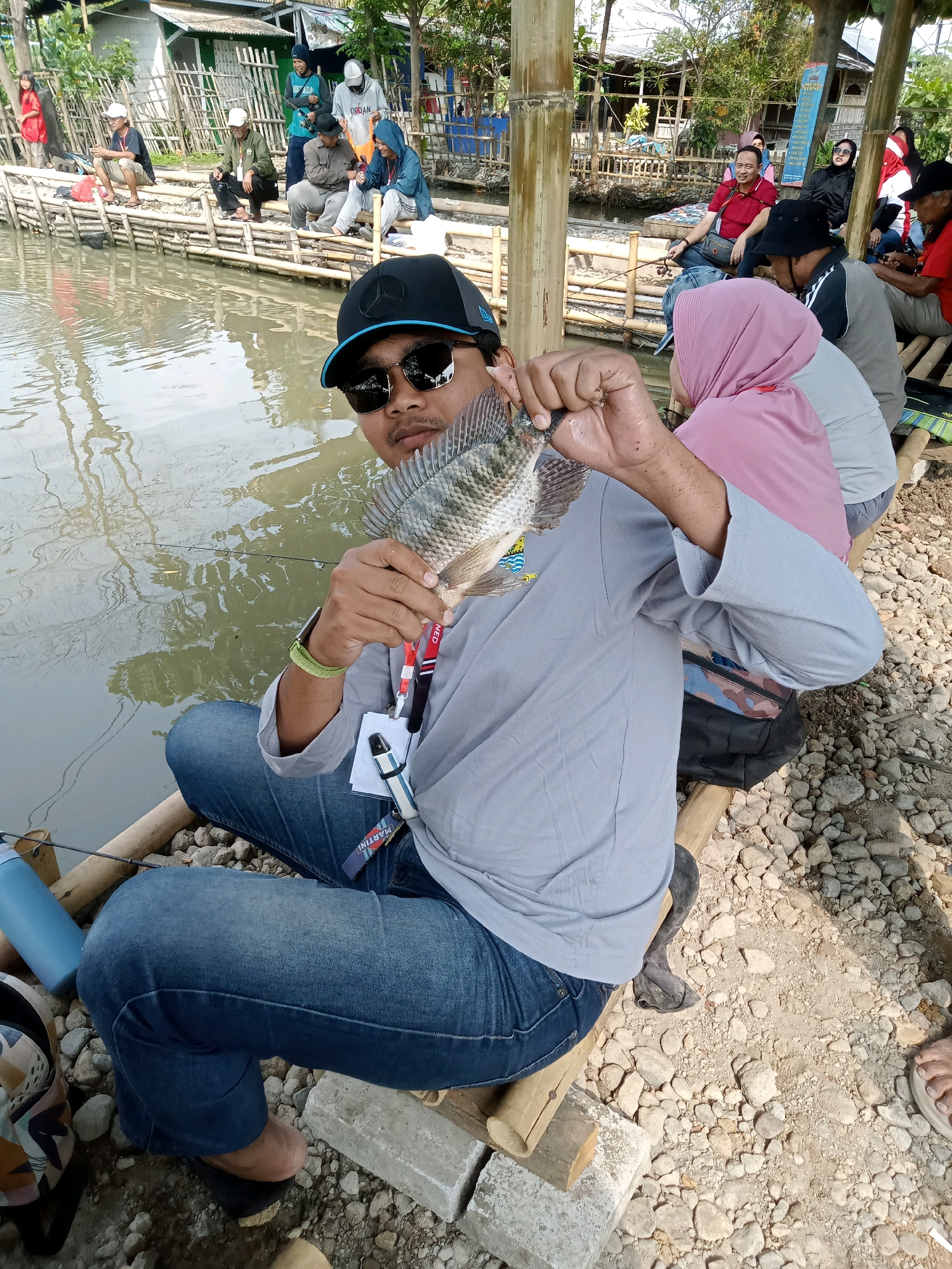 In the city of Kecomberan, Indonesia, on a bright sunny day, a group of people are engaged in fishing activities along the Kecomberan River. The foreground is dominated by a man sitting with his legs crossed, holding a freshly caught fish in his right hand. He is wearing a black baseball cap, sunglasses, a grey long-sleeved shirt, and blue jeans. The fish, a tilapia, is held up for display. Around him, other individuals are also seated on a wooden structure adjacent to the river, some with fishing rods. The riverbank is lined with bamboo poles and some houses are visible in the background. The ground is covered with small stones and dirt. The overall scene depicts a recreational fishing outing in a rural Indonesian setting.