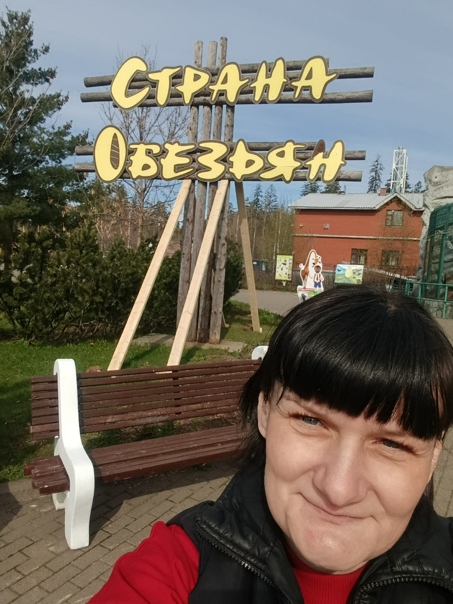 A woman takes a selfie at what appears to be an outdoor attraction, likely in Izhevsk, Russia. In the foreground, the woman with dark hair and bangs, wearing a red shirt under a black jacket, smiles or smirks directly at the camera. Behind her, a prominent rustic wooden sign features large yellow Cyrillic text that reads "СТРАНА ОБЕЗЬЯН" (Strana Obezyan), translating to "Monkey Country" or "Land of Monkeys," a known attraction in Izhevsk.
To the left of the sign, mature coniferous trees are visible, while sparse, bare deciduous trees suggest it might be spring or late autumn. A wooden park bench with distinctive white-painted supports is positioned in the lower-left corner of the image. In the mid-ground, beyond the sign, a red brick building with a gray roof and several informational displays or character cutouts can be seen. Further back, a white metal structure, possibly a tower or part of a ride, rises against a clear blue sky. The setting appears to be a bright, sunny day, with ample light illuminating the scene. The woman's expression conveys happiness or amusement.