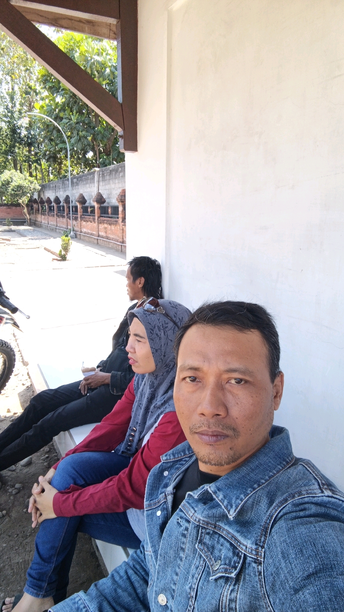 Three people are sitting on a white bench outdoors. The person in the foreground is a man with short dark hair and a goatee, wearing a denim jacket and a black t-shirt. He is looking at the camera, with his right arm resting on his lap. To his left, a woman wearing a grey hijab and a red top is sitting with her hands clasped in her lap. Behind her, a man with dark, tousled hair is looking to his left. A motorcycle is partially visible to the left of the frame. The background shows a white wall, trees, a fence made of brick and ornamental posts, and a paved area. The scene appears to be in Kendalbulur, Indonesia.