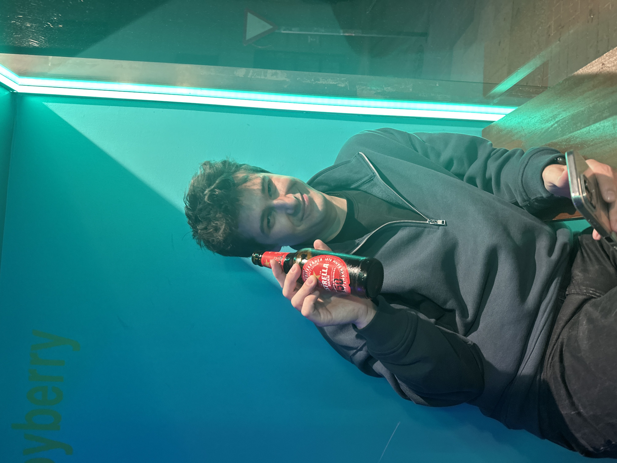 A man is sitting at a cyan wall and holding a bottle of Estrella Damm beer. The man is wearing a dark hoodie and dark jeans. He has short dark hair and is smiling at the camera. The wall is a cyan color with the word "cyberberry" written on it. The man is sitting on what appears to be a bench with an edge near the top. The beer bottle is red with gold lettering. The location is suggested to be Madrid, Spain, due to the presence of Estrella Damm beer.