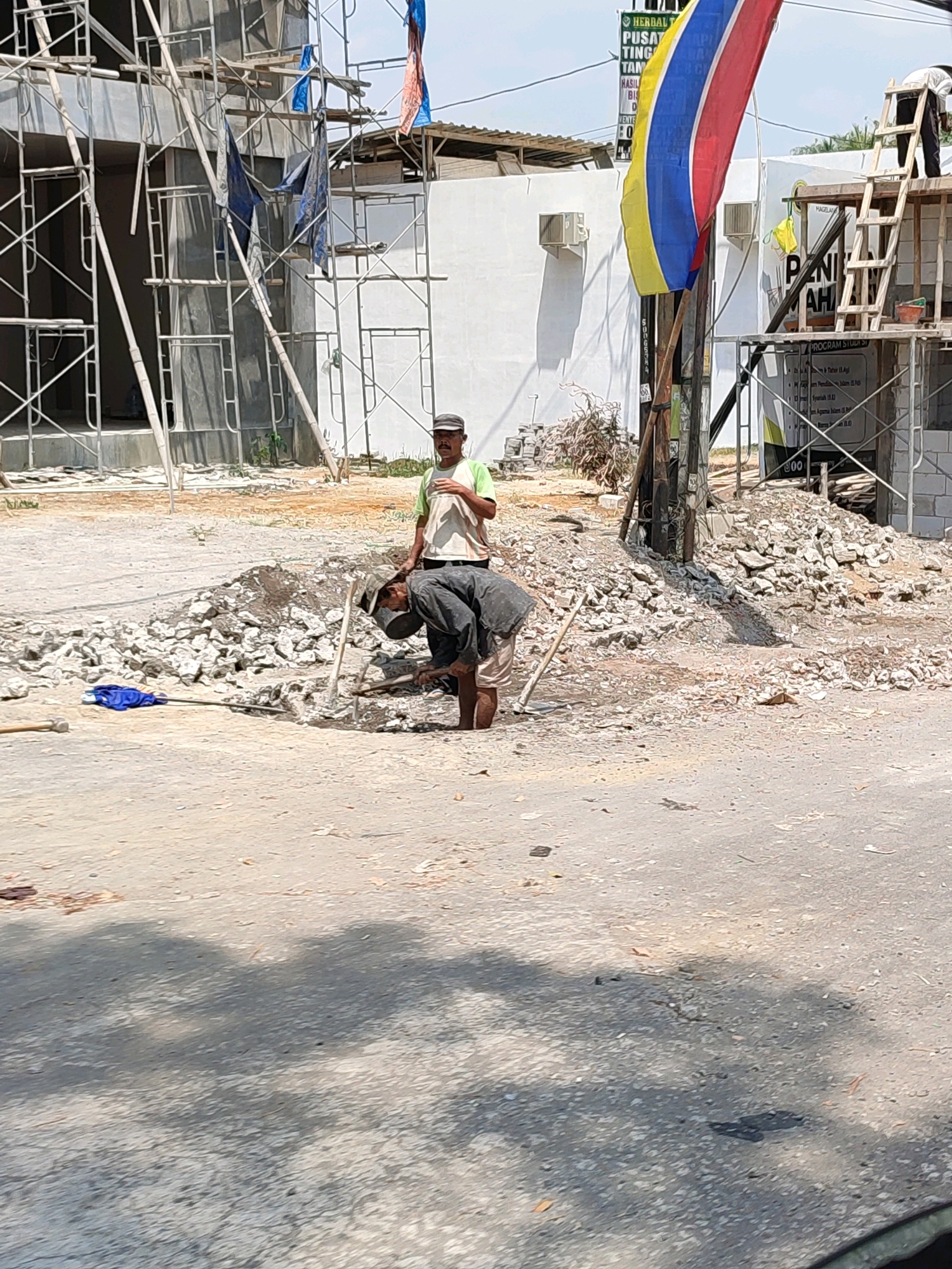 Two men are working at a construction site in Tanggulrejo, Indonesia. One man, wearing a dark shirt and light-colored pants, is bent over and appears to be digging or breaking up concrete with a shovel. He is positioned in front of a pile of rubble. The second man, wearing a light-colored t-shirt and a dark cap, stands upright behind the first man, observing. He is facing towards the man working.
In the background, a building under construction is visible, supported by scaffolding. There is a large flag with yellow, red, and blue stripes flying from a pole. Construction materials, including bricks and debris, are scattered around the site. The ground is a mix of dirt and pavement. The overall environment suggests an active construction project in progress under daylight conditions.
