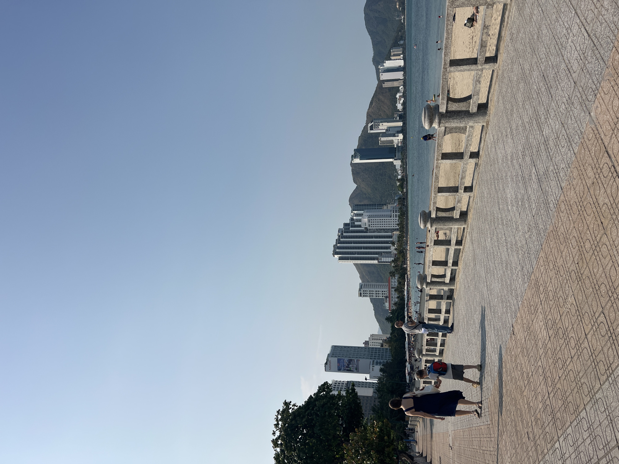The image captures a scene on a seaside promenade in an urban area. The sky is a clear blue. A row of modern, high-rise buildings lines the shore against a backdrop of sloping, green mountains. The beach is visible, situated beside the water. On the right side of the frame is a paved walkway with a patterned surface. Along the walkway are concrete structures that appear to be either decorative or serve as a low barrier. Several people are present on the walkway, including a woman in a dark dress, a child with a red backpack, and another adult. These individuals appear to be walking together. There is also a tree visible in the lower-left corner of the frame.