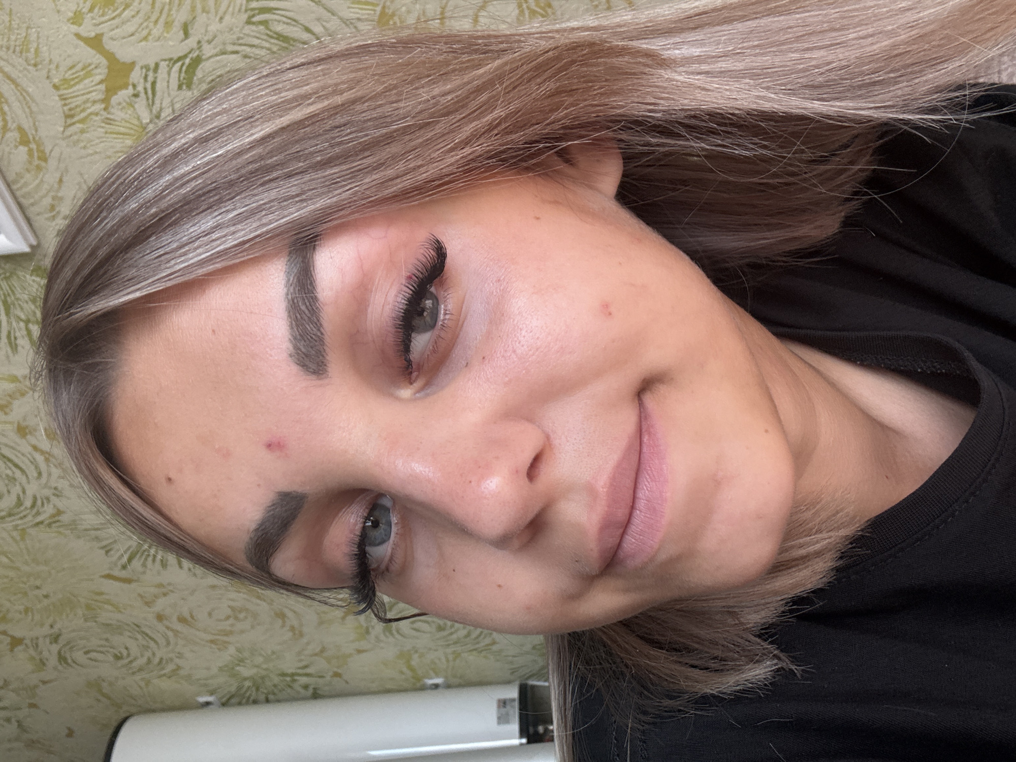 A woman with blonde hair and blue eyes is pictured from the chest up. She is wearing a black shirt and has on dramatic false eyelashes and well-defined eyebrows. She has a small red blemish on her forehead and a slight smile on her face. The background appears to be an indoor setting with a patterned wallpaper. The image does not contain any elements that would allow for the determination of the specific location within Krasnogorskoye, Russia.