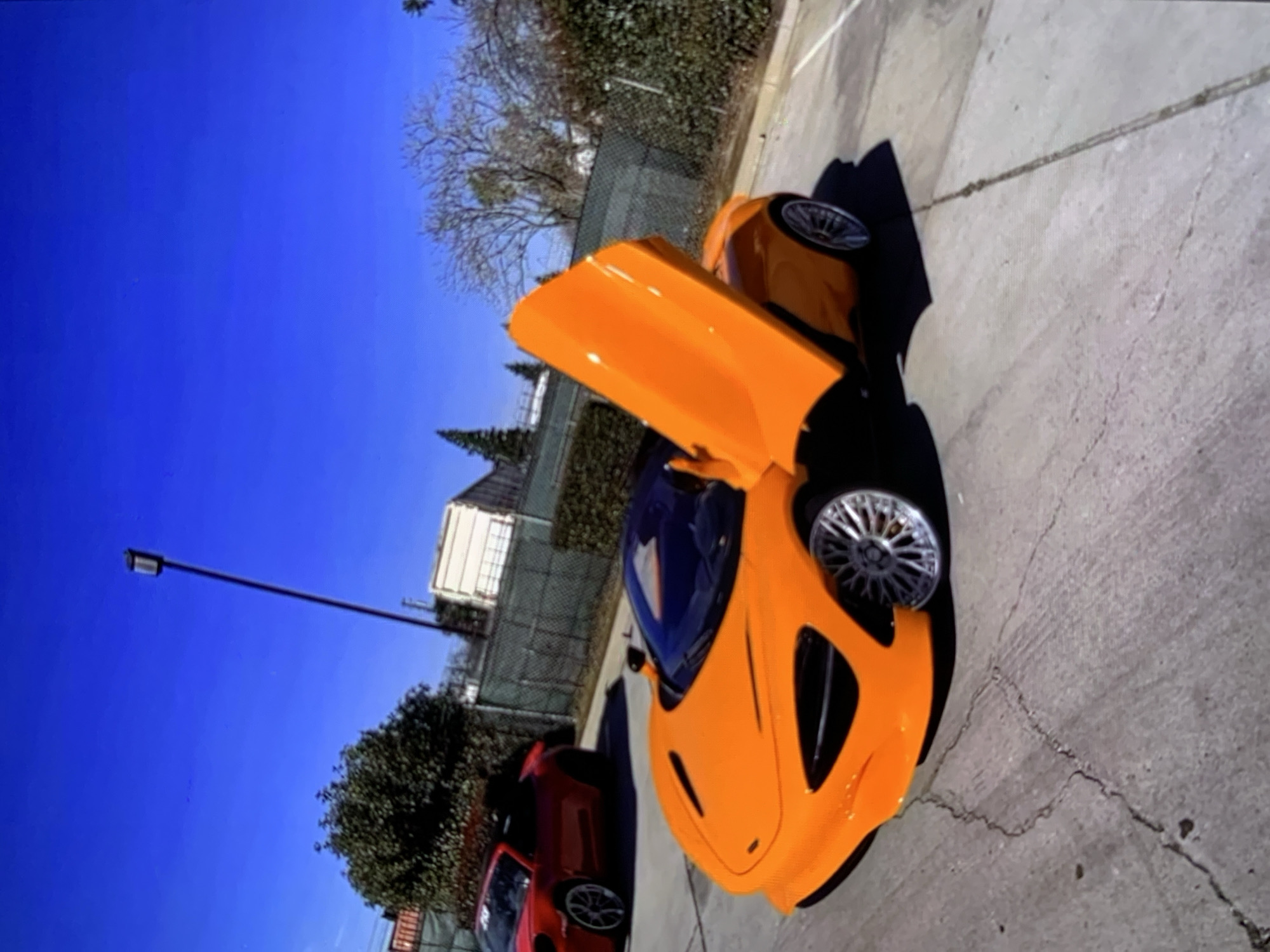 An orange McLaren 720S with its dihedral doors open is parked in a lot with a red Porsche 911 in the background. The McLaren's doors are pointed upwards and outwards from the vehicle, showcasing its interior and engine compartment. The car is parked on a cracked concrete surface, with tire marks visible. The environment appears to be an outdoor parking lot, with a fence and some vegetation in the background. The sky is clear and blue.