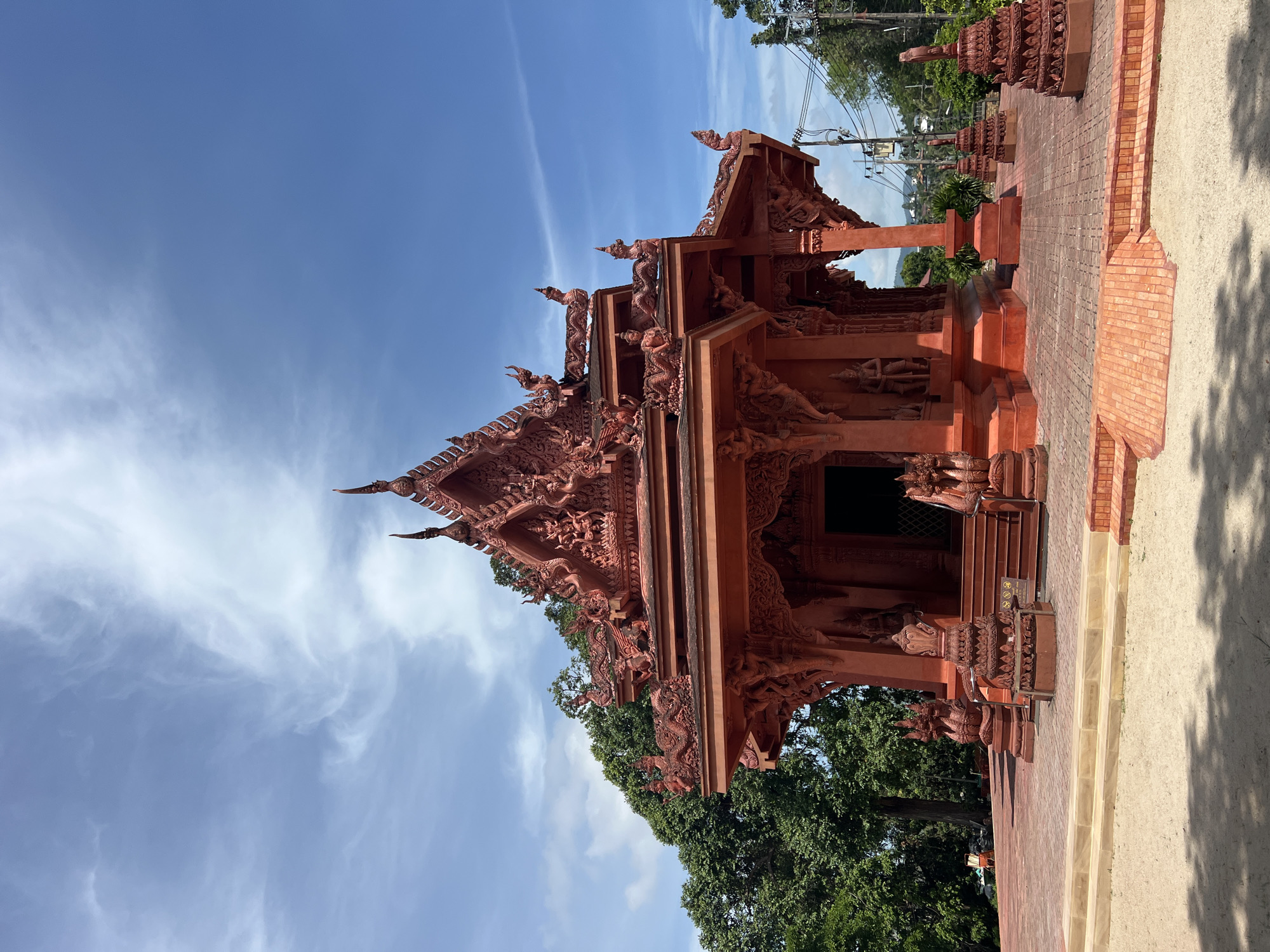 This image captures the exterior of a vibrant red Buddhist temple, likely the Wat Khao Phra Bat or a similar temple located in Baan Hua Thanon, Thailand. The temple features intricate architectural details, with ornate carvings and sculptures adorning its roofs and facade. The structure is a rich, deep red color, standing out against a bright blue sky dotted with wispy white clouds.
Surrounding the temple is a paved courtyard with red brick tiling. In the foreground, a sandy path casts dappled shadows from an unseen tree. To the left of the temple, lush green trees provide a natural backdrop, while to the right, power lines and distant foliage suggest an urban or semi-urban environment. Several smaller, decorative tiered structures, also red, are placed around the temple grounds. The overall scene is one of cultural and architectural significance, bathed in the bright light of day. No people are visible in the image.