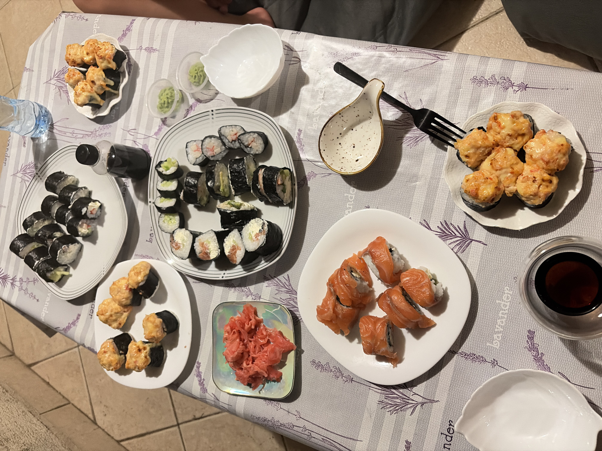 The image depicts a close-up, high-angle view of a table laden with various types of sushi and accompaniments, set on a lavender-patterned tablecloth. The scene appears to be indoors, possibly a dining area, given the tiled flooring visible in the upper left corner, which is characteristic of outdoor terraces or patios in coastal Mediterranean regions. While the image itself does not provide explicit clues to its location in Budva, Montenegro, the visual elements such as the tiled flooring, the casual dining setup, and the overall ambiance are consistent with a relaxed, potentially seaside establishment or a private residence in such a locale.
Several plates of sushi are arranged on the table. One plate contains several pieces of sushi with a creamy, orange topping, possibly baked salmon or spicy mayo. Another plate holds several rolls of sushi with visible cucumber and what appears to be crab stick, cut into bite-sized pieces. A third plate features several pieces of nigiri sushi topped with raw salmon. A small dish of pickled ginger, with its distinctive pink hue, is also present. There are also two small dishes containing wasabi, and a small white dipping bowl. A bottle of soy sauce is positioned near one of the sushi plates, alongside a clear glass bottle of water. A black fork rests on the table, positioned over one of the sushi plates. The lighting suggests it is daytime, possibly late afternoon given the warm tones and shadows. No people are directly visible in the frame, though the presence of a leg in the lower right corner suggests someone is seated at the table. The overall impression is a meal shared in a casual and inviting setting.