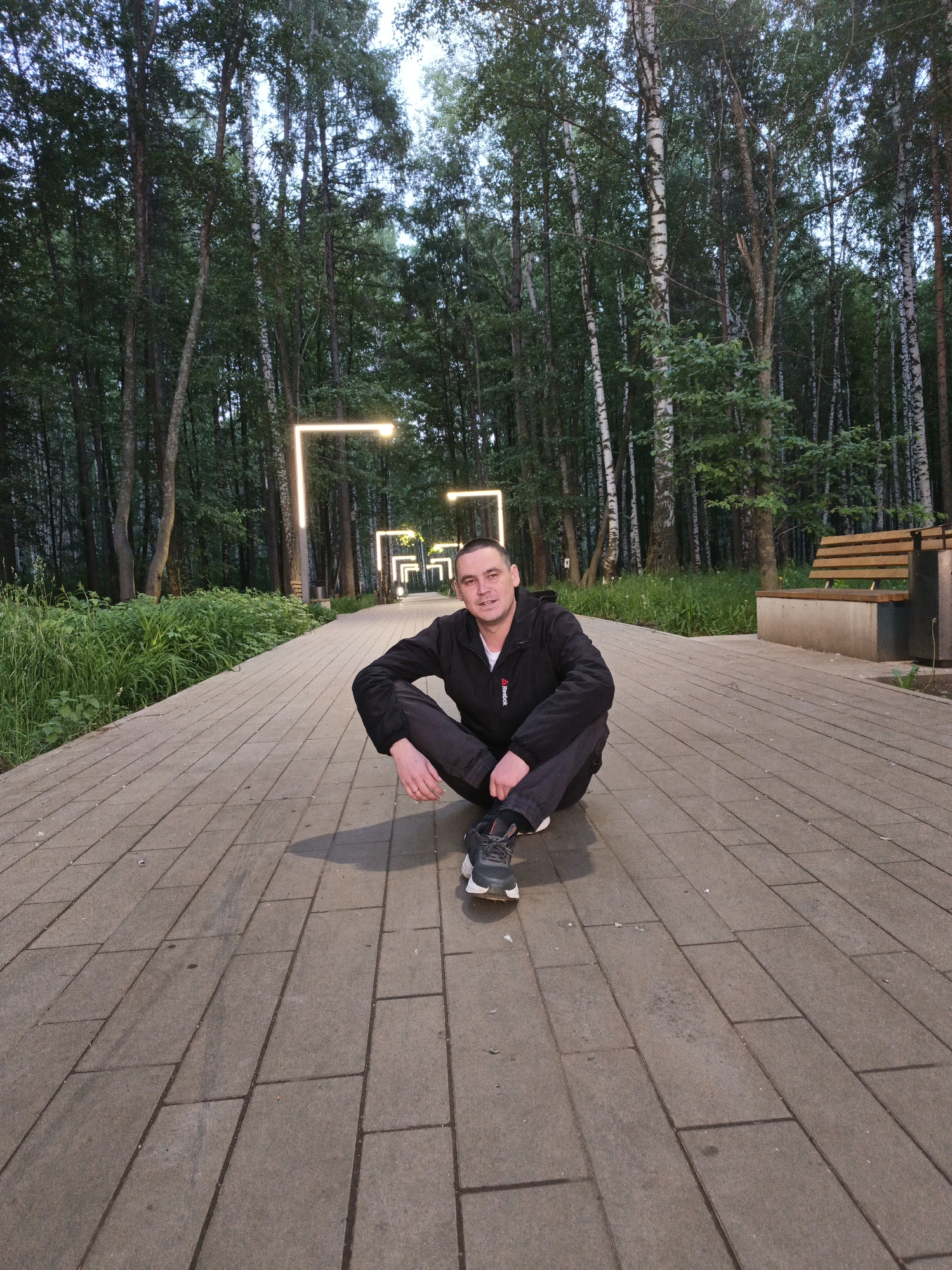 A man is sitting in a squatting position on a paved walkway in Chaikovsky, Russia. He is wearing a black jacket, dark pants, and sneakers. The walkway stretches into the distance, flanked by trees and illuminated by modern rectangular light fixtures. To the right of the man, there is a bench. The environment appears to be a park or recreational area.