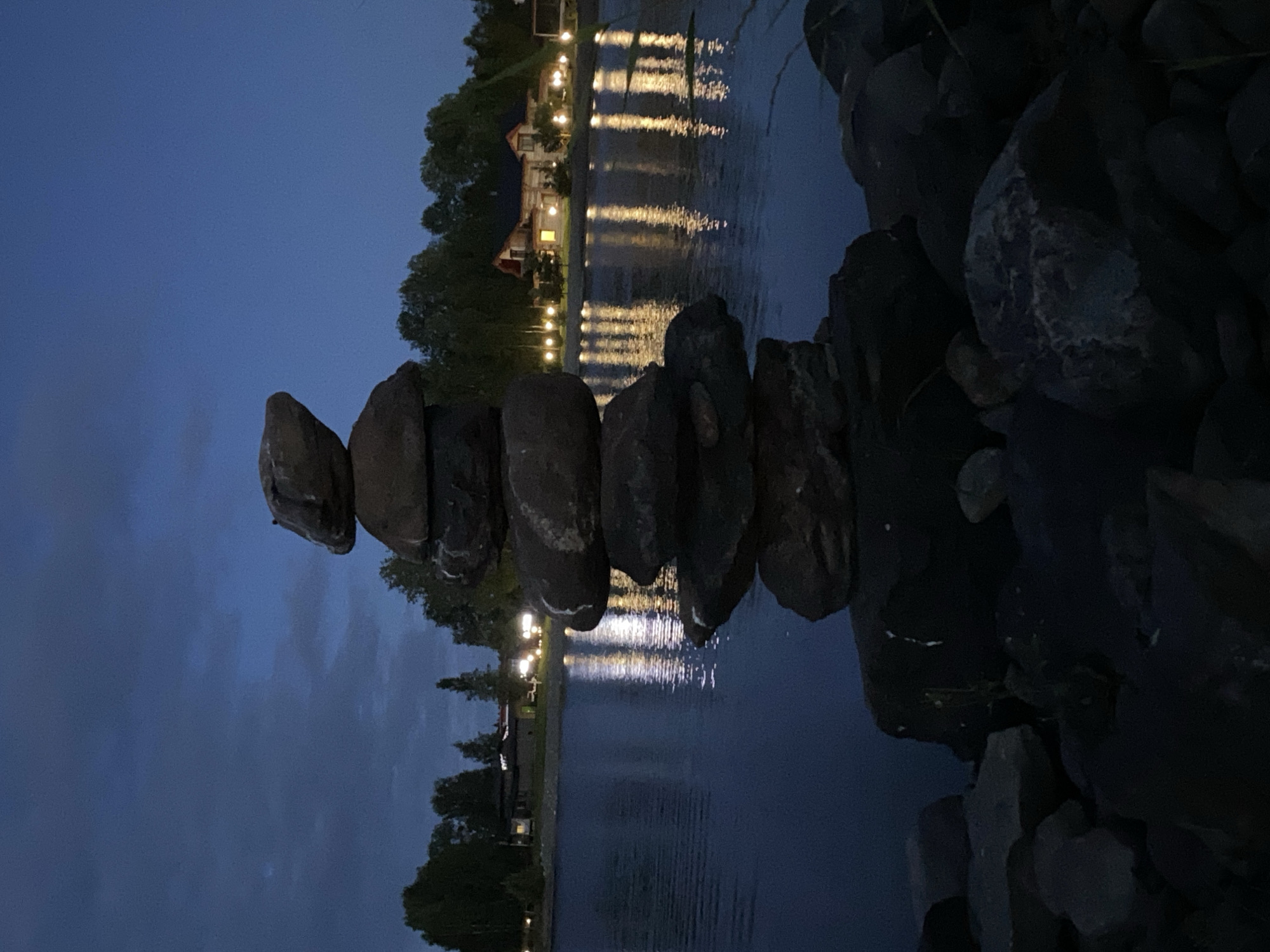 A cairn of stones is stacked precariously on a rocky shore at dusk. The sky is a deep twilight blue, with hints of clouds. A calm body of water, reflecting the lights of a nearby village, lies beyond the stones. The village, situated across the water, is characterized by houses with illuminated windows and a line of streetlights casting warm glows onto the water. Trees line the background, creating a silhouette against the dusky sky. The foreground consists of darker, rougher stones, partially obscuring the base of the cairn. The image was captured in Kirkkolakhti, Russia.