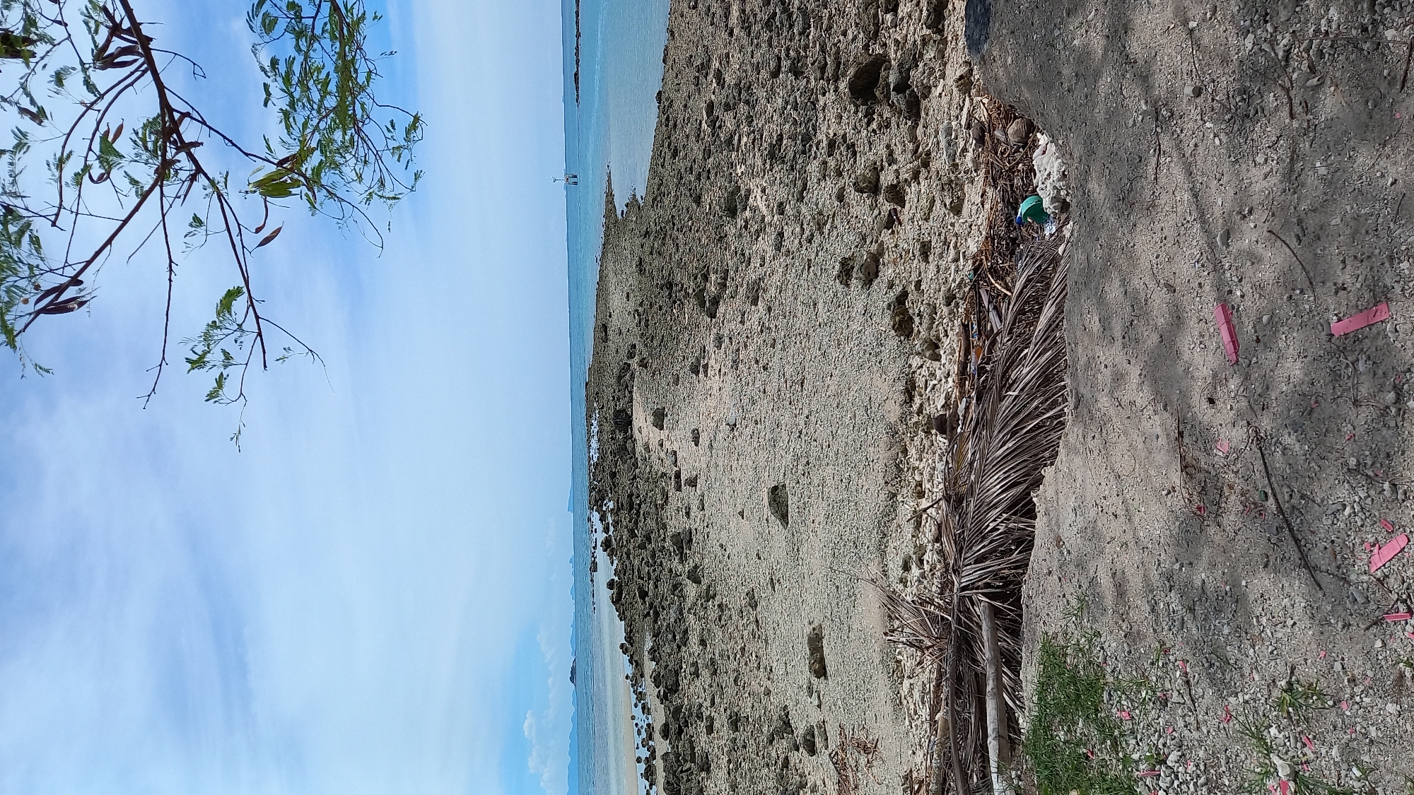 The image shows a rocky causeway extending into the ocean under a partly cloudy sky. The causeway is composed of gray and brown rocks, with some sandy patches and sparse vegetation. Dry palm fronds and debris are scattered on the ground in the foreground. In the distance, several islands are visible on the horizon. A small navigational marker stands in the water. The scene appears to be a coastal area in ตำบลเกาะพะงัน, Thailand.