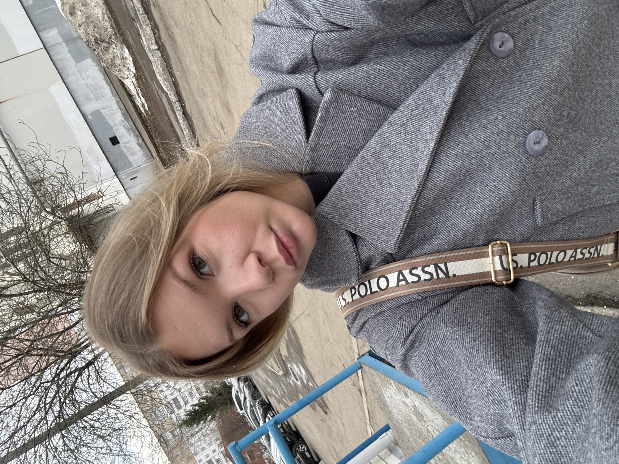 A woman with fair skin and light brown hair poses for a selfie outdoors, wearing a grey wool double-breasted coat. A U.S. Polo Assn. strap is visible across her body. The background shows a city landscape, including leafless trees, buildings, parked cars, and a blue metal railing. A patch of snow is visible near a building. Based on the general architectural style and context, the location is likely Perm, Russia.
