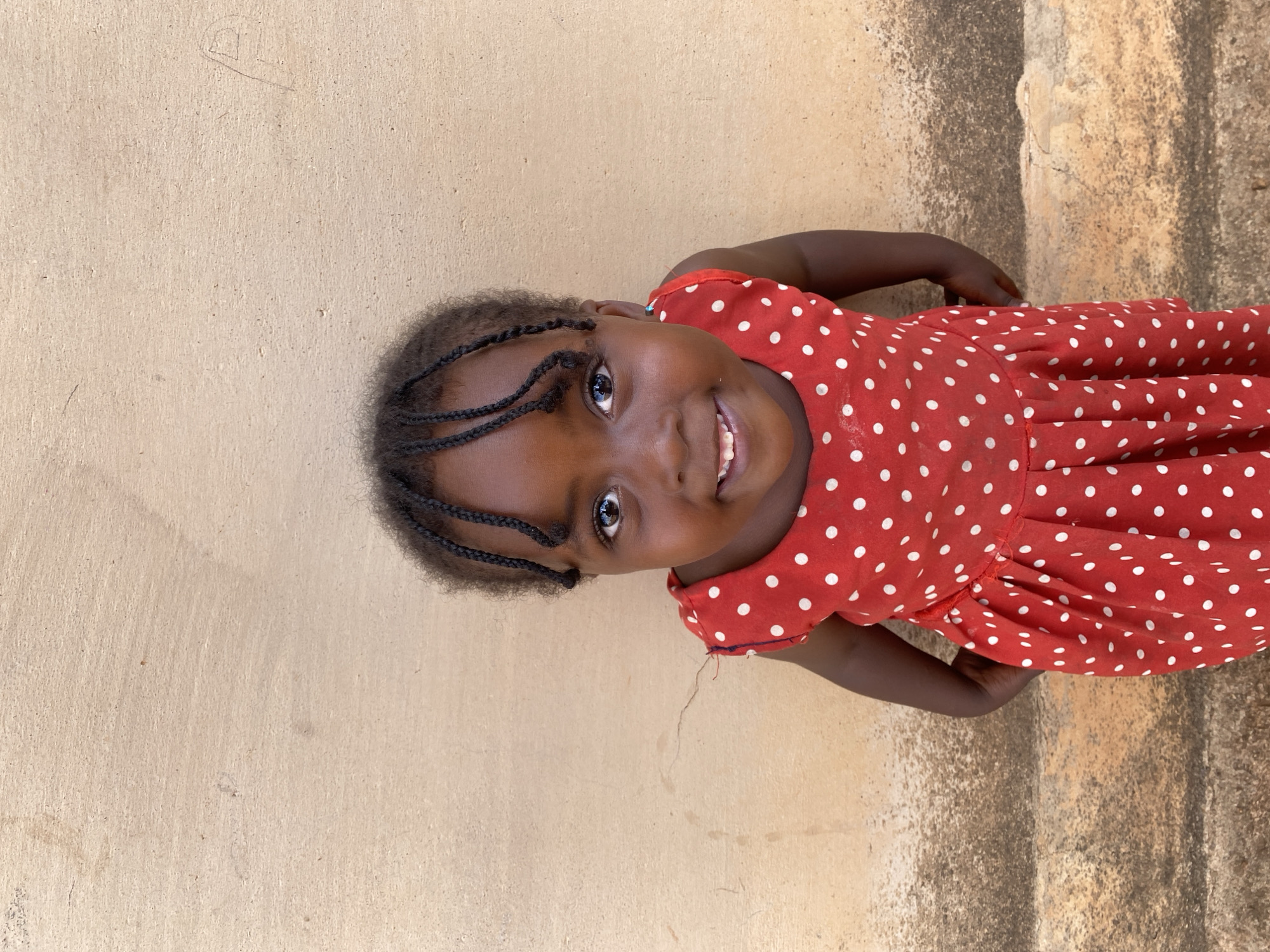 A portrait captures a young girl with dark skin, looking up and smiling directly at the camera. She wears a vibrant red dress adorned with numerous white polka dots. Her dark hair is styled in several small, neat braids that frame her face. The girl's eyes are wide and bright, reflecting light, and her smile is broad, revealing her teeth, conveying an emotion of joy and engagement.
The background consists of a plain, textured wall, predominantly light brown or beige in color. The wall shows subtle imperfections, varying textures, and some darker, possibly weathered patches. In the upper right area of the image, a faint, hand-drawn symbol or letter can be discerned on the wall. The scene is brightly lit, suggesting it was captured during the daytime. This media file originates from Kuru Dungus, Nigeria.