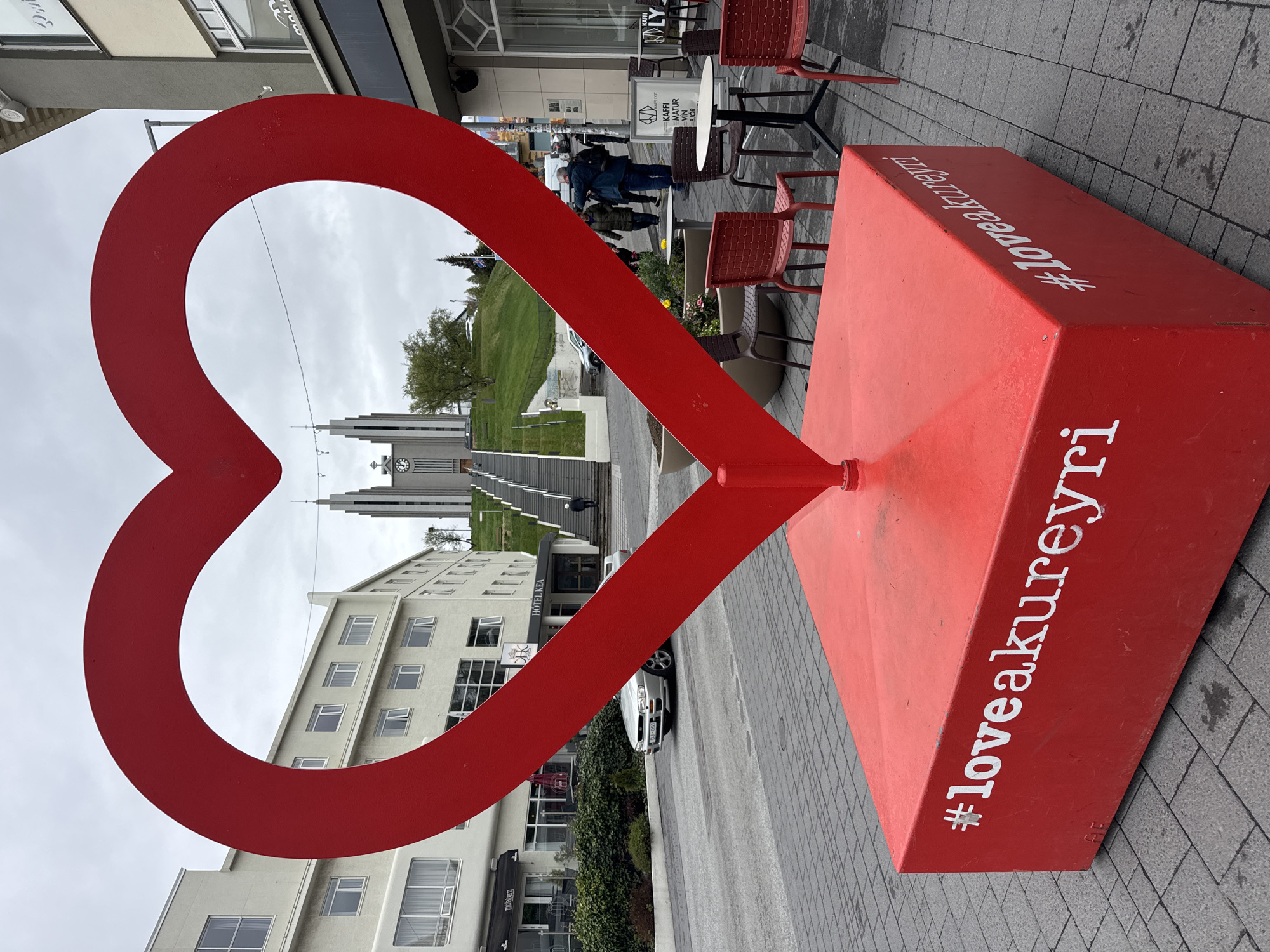 This image captures an outdoor urban scene in Akureyri, Iceland, characterized by a large, bright red heart sculpture in the foreground. The sculpture, serving as a prominent landmark, stands on a rectangular base that features the white text "#loveakureyri" along its side.
Through the heart's opening, the iconic Akureyri Church (Akureyrarkirkja) is visible in the background, distinguished by its tall, angular twin towers and a long set of steps leading up a hillside. A person is seen ascending these steps. To the left of the church, a multi-story building displays the "HOTEL KEA" sign.
On the right side of the frame, several red chairs and small tables are arranged on a paved sidewalk, indicating an outdoor cafe or restaurant. A partial sign near this area reads "Kaffi og Vín". A few people are present in this area, either walking or patronizing the establishment. A silver car is parked on the street, and a lush green hillside with trees adds natural elements to the urban landscape.
The weather appears overcast, with a cloudy sky providing diffused lighting across the scene, suggesting a daytime shot. The overall impression is one of a vibrant town center, with the heart sculpture celebrating local affection for Akureyri.