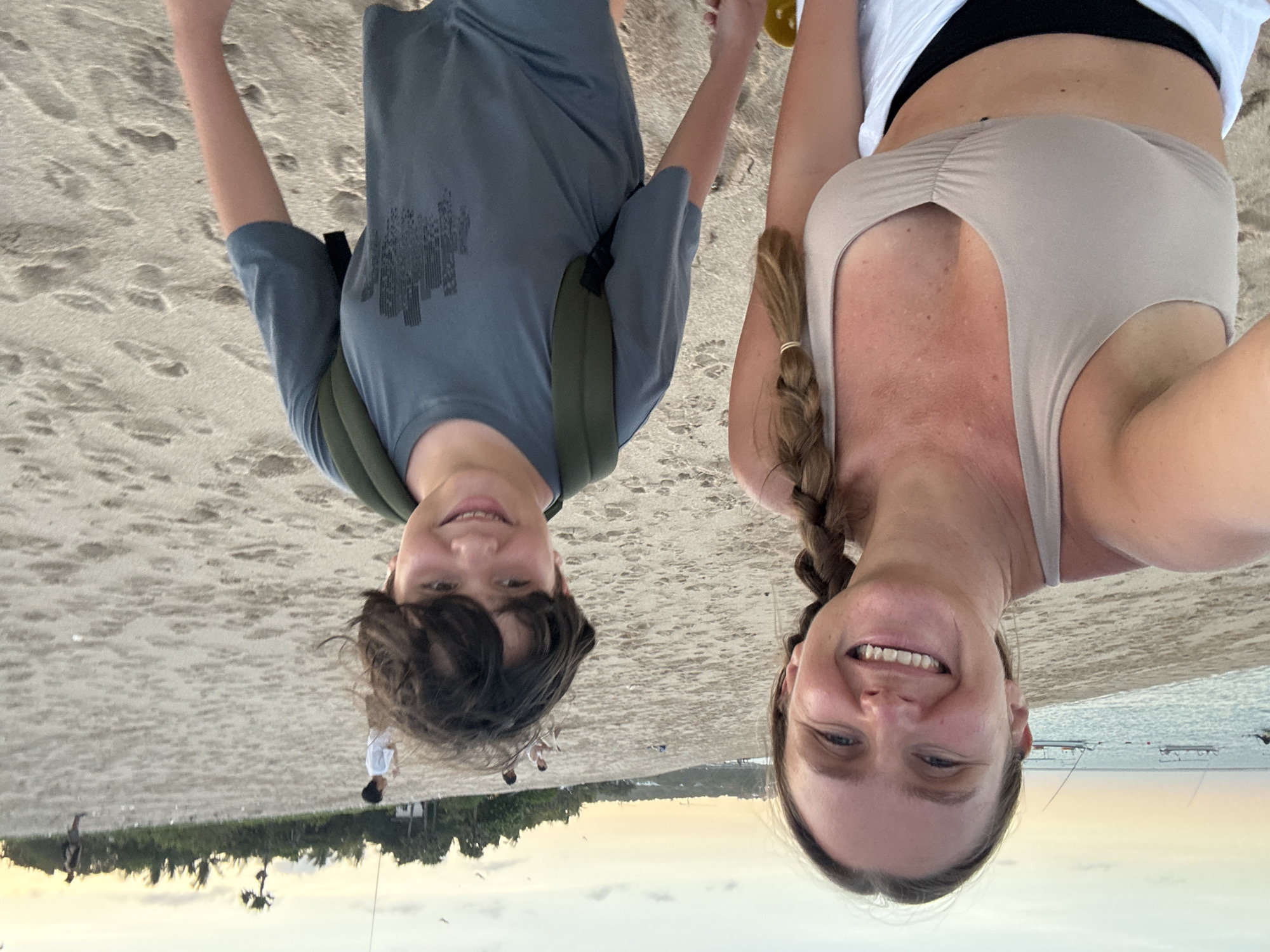 A woman and a boy are posing upside down on a sandy beach. The woman has blonde hair styled in a braid and is wearing a light-colored tank top and a white shirt over a black top. The boy has brown hair and is wearing a gray t-shirt with a dark-colored graphic and a green backpack. The background shows a beach scene with several people in the distance, trees and buildings along the shoreline, and boats on the water. The sky is visible near the horizon, suggesting it may be sunset or sunrise. Based on the location, it is likely they are on a beach in Kuta Selatan, Indonesia.