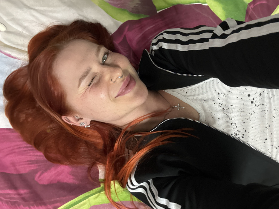 A woman with prominent red hair and freckles is depicted lying on patterned bedding in Rostov-on-Don, Russia. She is smiling broadly and winking with her right eye, gazing directly towards the camera. She wears a black athletic jacket with white stripes on the sleeves, partially unzipped over a white ribbed tank top with black speckle patterns. A silver-colored cross pendant hangs from a chain around her neck. Her visible right ear features multiple silver-colored piercings, including a butterfly or flower-shaped stud on the lobe and other studs on the helix and tragus. A silver-colored hoop nose ring is present in her left nostril. Her reddish-orange hair is spread out around her head on the fabric background, which displays a large, abstract floral pattern in shades of pink, purple, and green. The image is a close-up, taken from an overhead perspective.