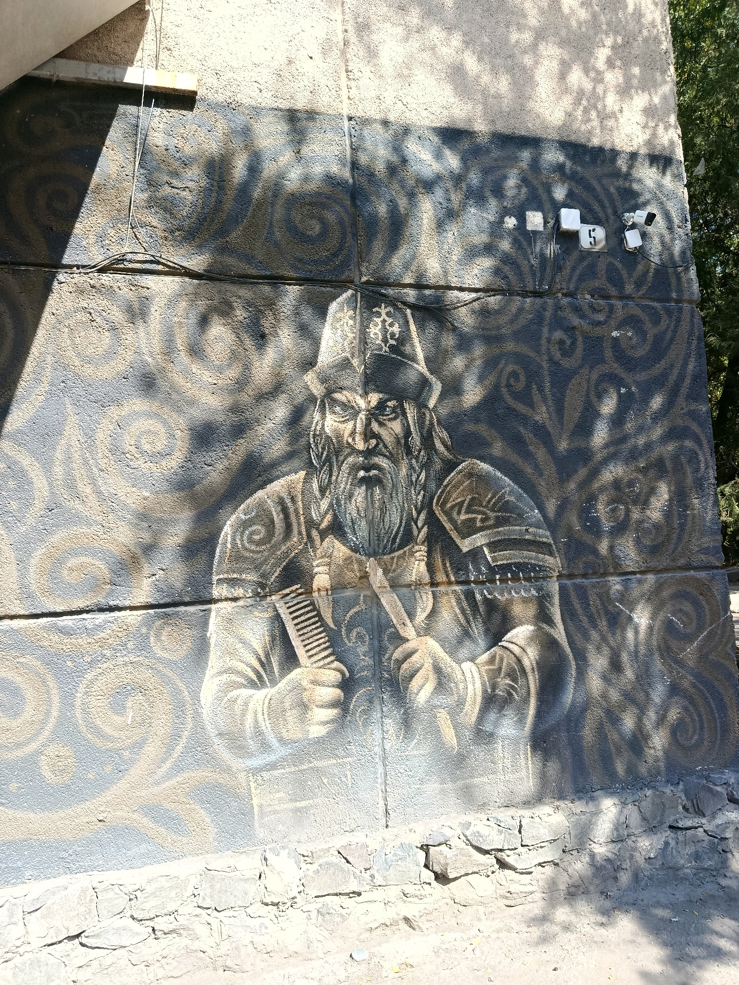 This image depicts a mural of a warrior, possibly a historical figure, painted on the exterior wall of a building in Bishkek City, Kyrgyzstan. The mural features a man with a long, braided beard, wearing a traditional hat and armor. He is holding a comb-like object in his hands. The background of the mural is decorated with swirling patterns in dark blue and beige tones. The wall itself appears to be textured and has some signs of wear.
The mural is located on a building with a light-colored facade above it. Electrical wires and what appear to be outdoor electrical outlets and security cameras are visible on the wall, to the right of the mural. The lower part of the building's exterior is constructed with rough-hewn stones. Sunlight casts shadows on the wall, creating a play of light and dark across the mural and the building's facade. Trees and foliage are visible in the background on the right side of the image. The ground in front of the building appears to be dirt or gravel.