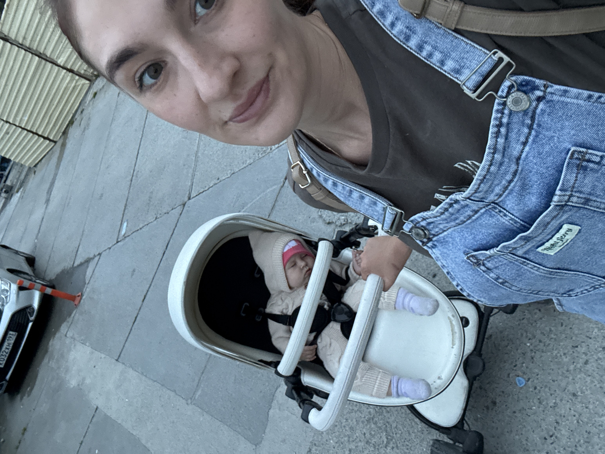 A woman takes a selfie with a baby in a stroller on a paved sidewalk. The woman wears denim overalls over a brown t-shirt. The baby is bundled in a knitted outfit with a hat. A car is partially visible on the left side of the frame. The license plate of the car reads "K032XH 193." This image was likely taken in Sochi, Russia, given the license plate number.