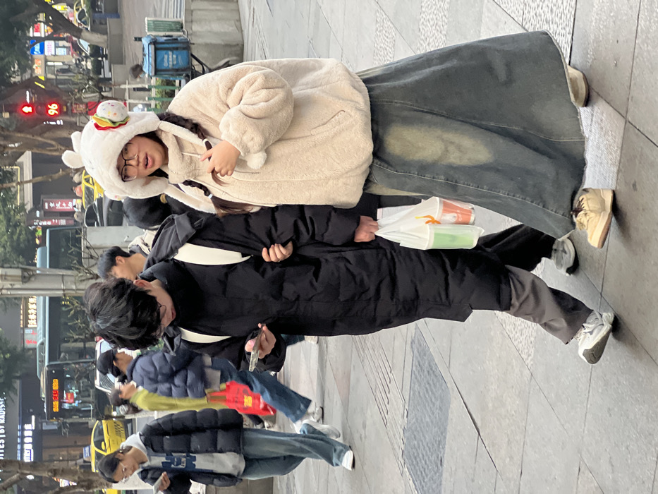 Two young adults of East Asian descent walk arm-in-arm on a paved city sidewalk in Yuzhong District, China. The male subject on the left wears a long black puffer jacket, dark pants, and white athletic shoes, with a white backpack strap visible. He wears glasses, has short dark hair, and looks down at a smartphone in his left hand. In his right hand, he carries a clear plastic bag containing one green and one orange/brown cup. The female subject on the right wears a cream-colored fuzzy zippered jacket, a long faded blue denim skirt, and light-colored platform shoes. She wears glasses and a white fuzzy hat with animal ears and a small hamburger ornament on its left side; two dark braids extend from beneath the hat. Her mouth is slightly open, and she looks forward or slightly right.
The background shows a busy urban street with vehicular traffic, including yellow taxis and a blue city bus. A red traffic light with a digital countdown displaying "96" is visible. Storefronts with Chinese characters and partial English text ("BY MADIS") line the street. Additional pedestrians are present in the mid-ground, some looking at smartphones. The sky appears overcast. A blue utility box is positioned on the right side of the frame.