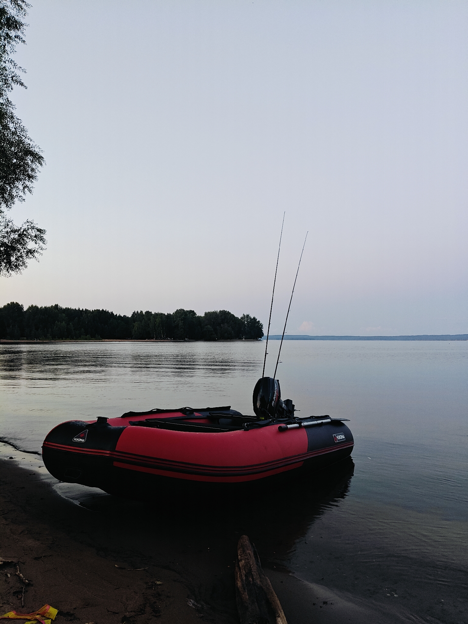 A red and black inflatable boat is pulled up onto a sandy shore. Two fishing rods are secured in the boat's stern, with an outboard motor visible behind them. The boat is on the edge of a calm body of water. In the background, a dense forest lines the distant shore. The sky is a pale, overcast gray, suggesting either early morning or late evening. The overall scene is tranquil and rural, consistent with a location like Noviy, Russia, which is known for its natural landscapes.