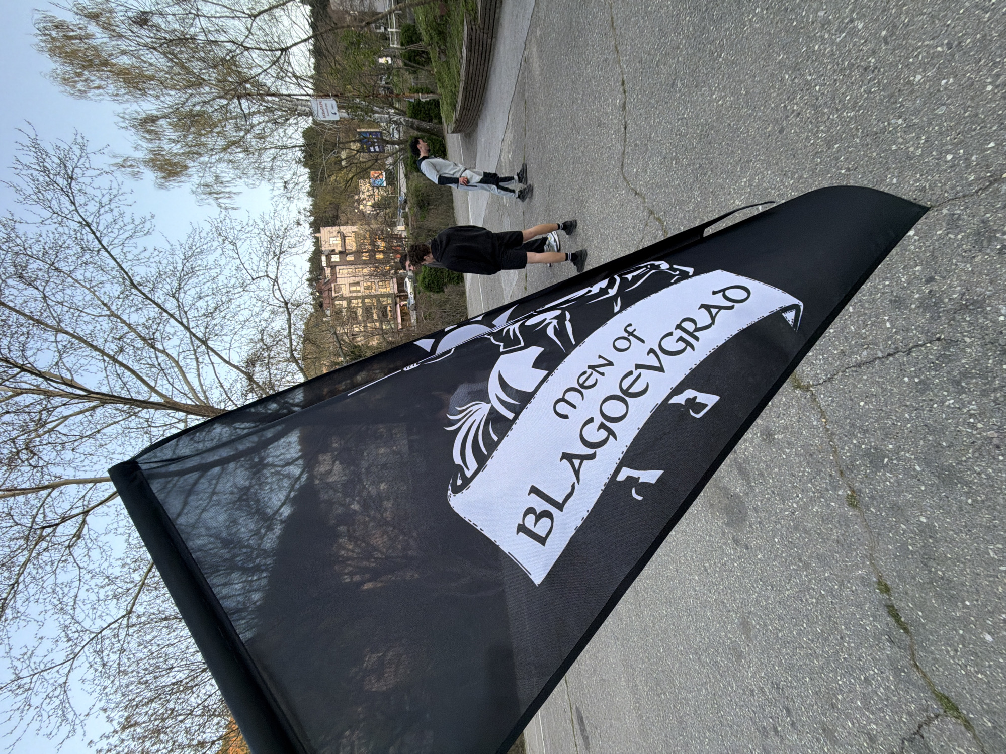 The image shows a black flag with white text that reads "Men of Blagoevgrad" in an outdoor setting. Two individuals are visible in the background; one is wearing a grey tracksuit and the other is wearing a black hoodie and shorts. The flag is positioned in the foreground, partially obscuring the background. The setting appears to be a paved area, possibly a sidewalk or courtyard, in the city of Blagoevgrad, Bulgaria. Buildings are visible in the background, along with bare trees. The sky is light blue. The image is oriented at an angle.