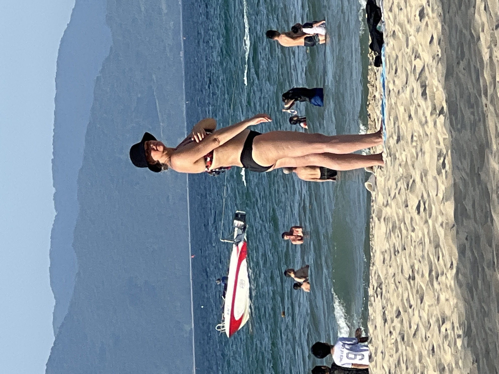 A woman in a black wide-brimmed hat and a bikini stands on a sandy beach facing the ocean in Da Nang, Vietnam. Her arms are crossed over her chest. Behind her, the blue ocean is dotted with several people wading in the water. A white and red boat is visible in the distance, moving away from the shore. Mountains form the background, shrouded in a light haze. The beach is covered in light-colored sand, with some shadows cast across it.