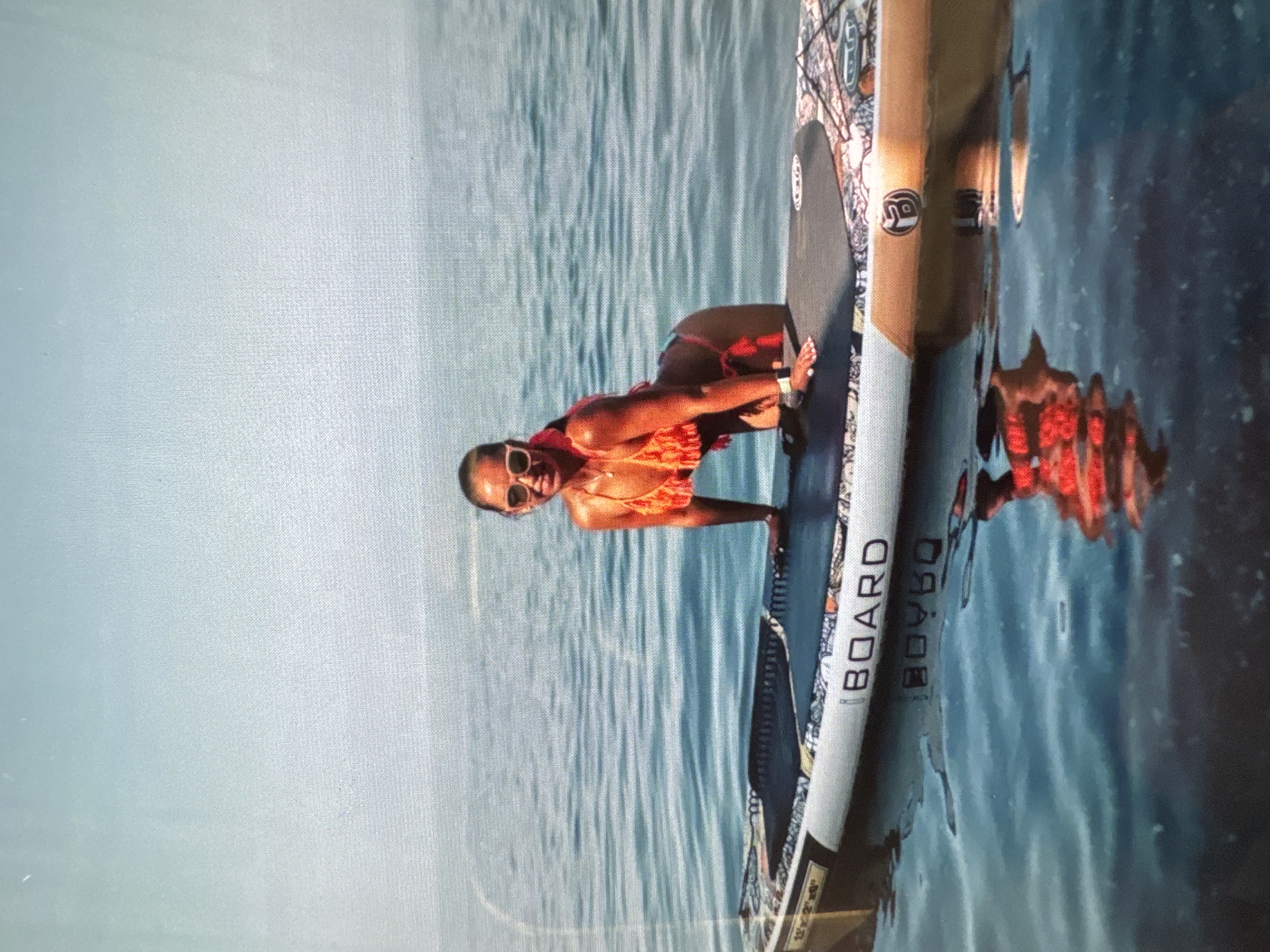 A woman in an orange bikini is posing on a paddleboard in the water. She is wearing sunglasses and her hair is pulled back. The paddleboard has a decorative pattern on the side, and the words "BOARD" and "DRAGON" are visible in text. The water is calm and blue, reflecting the sky and the paddleboard. The scene appears to be a sunny day, likely in the afternoon, given the lighting. The woman's pose suggests a relaxed and confident demeanor. The setting is a body of water, possibly the sea, given the hint of Sochi, Russia as a location.
