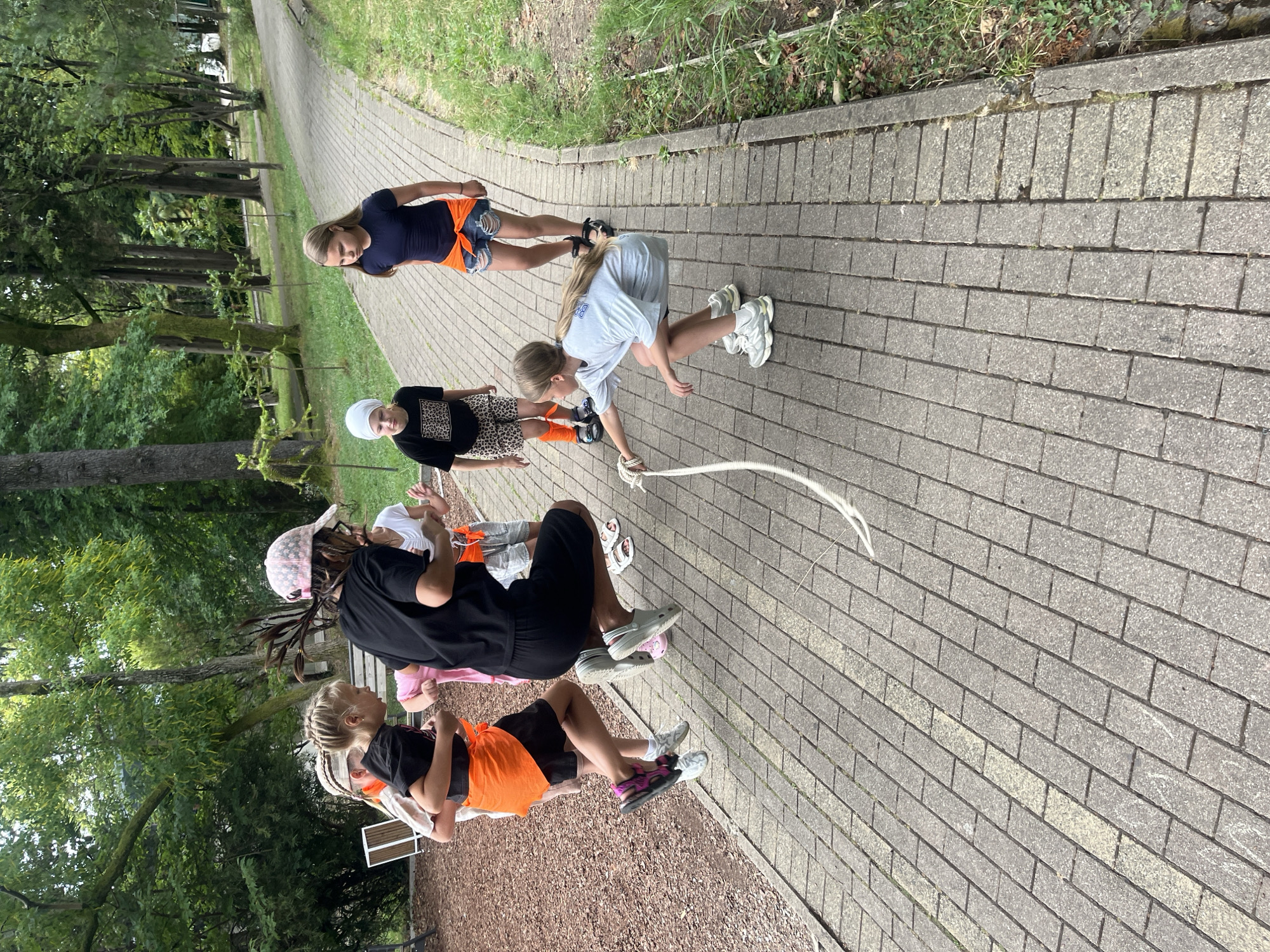 In a park setting, likely in Sochi, Russia, a group of children are engaged in a game of jump rope. A young girl in a light blue t-shirt and dark shorts is in the foreground, crouched low and holding one end of a thick, white rope. She appears to be initiating the jump for another child. In the mid-ground, a girl in a black t-shirt and pink and white baseball cap is mid-air, jumping over the rope. She wears dark shorts and a bright orange sash tied around her waist. To her left, two other children in similar attire, with orange sashes and dark shorts, are also jumping or preparing to jump. Further back on the paved path, two more girls are observing or participating. One, wearing a dark t-shirt and ripped denim shorts with an orange sash, stands facing away. Another, with a white headscarf and a black and leopard print t-shirt, is looking towards the jump rope action. The environment is characterized by a paved walkway lined with grass and trees, suggesting a public park or recreational area. The overall scene depicts children actively playing outdoors.
