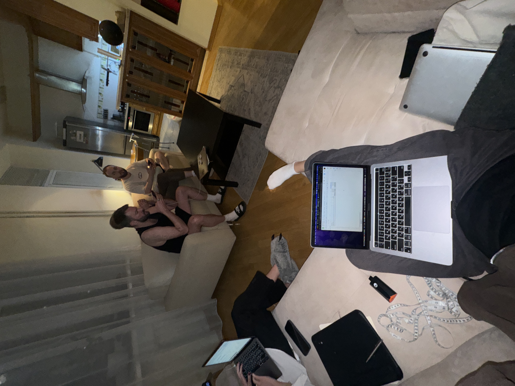 A slightly elevated, tilted aerial shot depicts an interior scene with multiple individuals and technological devices. A kitchen area with stainless steel appliances is visible in the background, along with wooden cabinetry.
Two men are seated on a light-colored sofa. One of them is wearing a light-colored shirt and dark pants, and is using a handheld device. The other is wearing a tank top and dark shorts.
In the foreground, a person, partially visible, is seated on a white sofa, wearing dark sweatpants and grey slippers. They are using a silver laptop. Next to the laptop, there's a black object with an orange tip, what appears to be a tablet, and a measuring tape.
Another person is visible further down the sofa using another laptop.
The room is illuminated and appears to be in a residential setting. There is a black table between the sofas with a silver laptop laying on it. The location of the image is not provided or evidenced in this image.