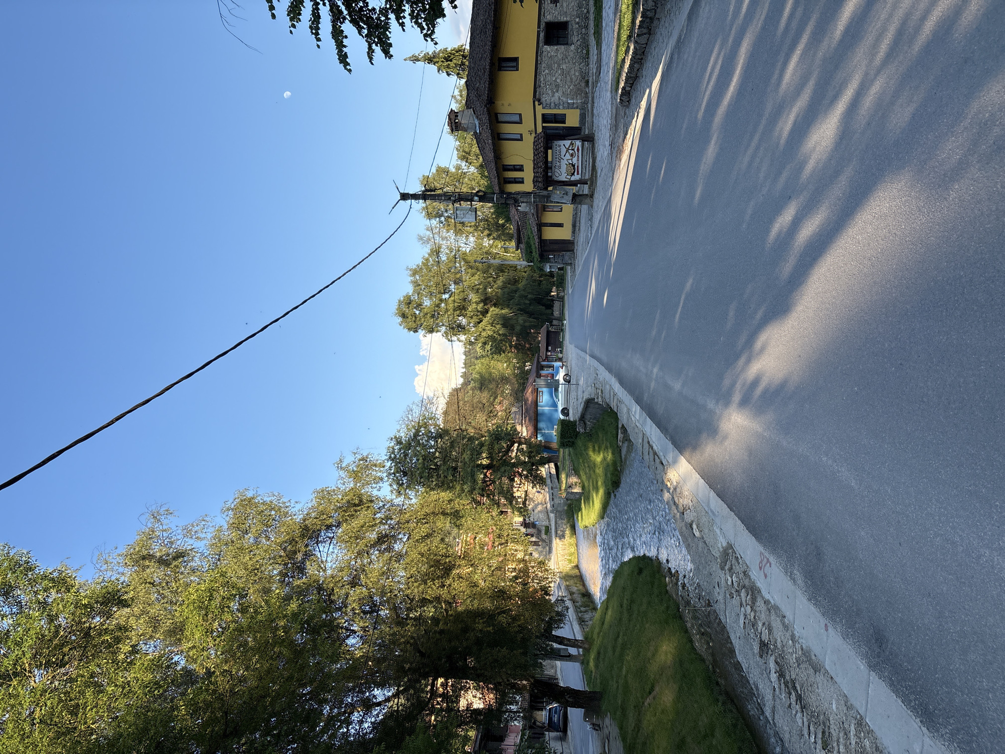 The image depicts a street scene in Koprivshtitsa, Bulgaria. A paved road runs diagonally across the frame, bordered by a stone wall and a stream. Lush green trees line the other side of the road, casting dappled shadows on the asphalt. A two-story building with a yellow facade and a tiled roof is visible, featuring several windows and a stone foundation. A sign with text and graphics, possibly indicating a restaurant or establishment, is mounted near the entrance of the building. The sky is clear and blue, with the faint presence of the moon. A power line stretches across the upper left portion of the image. The overall scene suggests a tranquil daytime setting in a picturesque town. The lighting indicates it is likely late afternoon or early morning, given the long shadows.